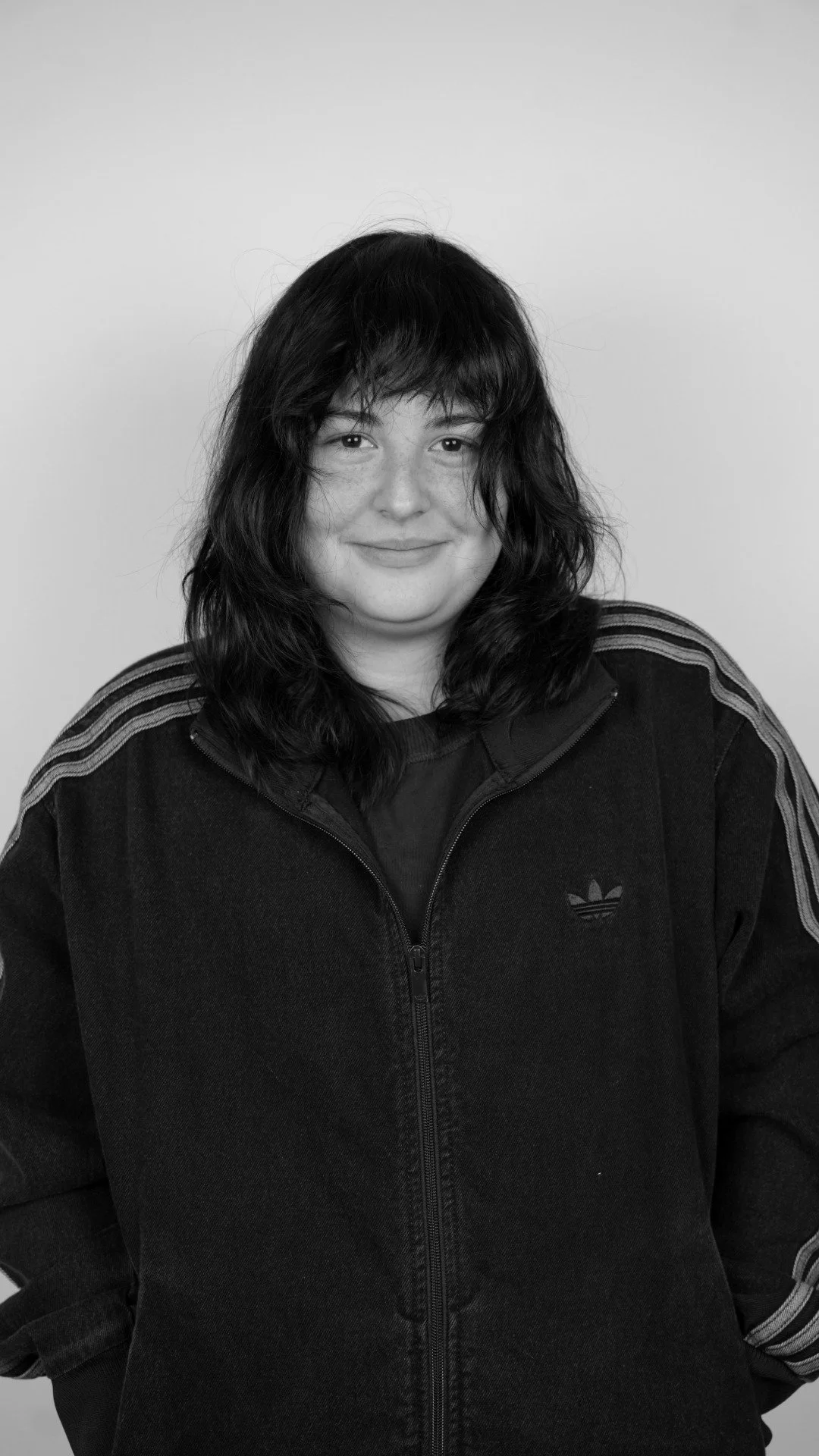 Black and white portrait of a woman with shoulder-length wavy hair, wearing a black Adidas zip-up jacket with stripes on the sleeves and the Adidas logo on the chest, smiling gently.
