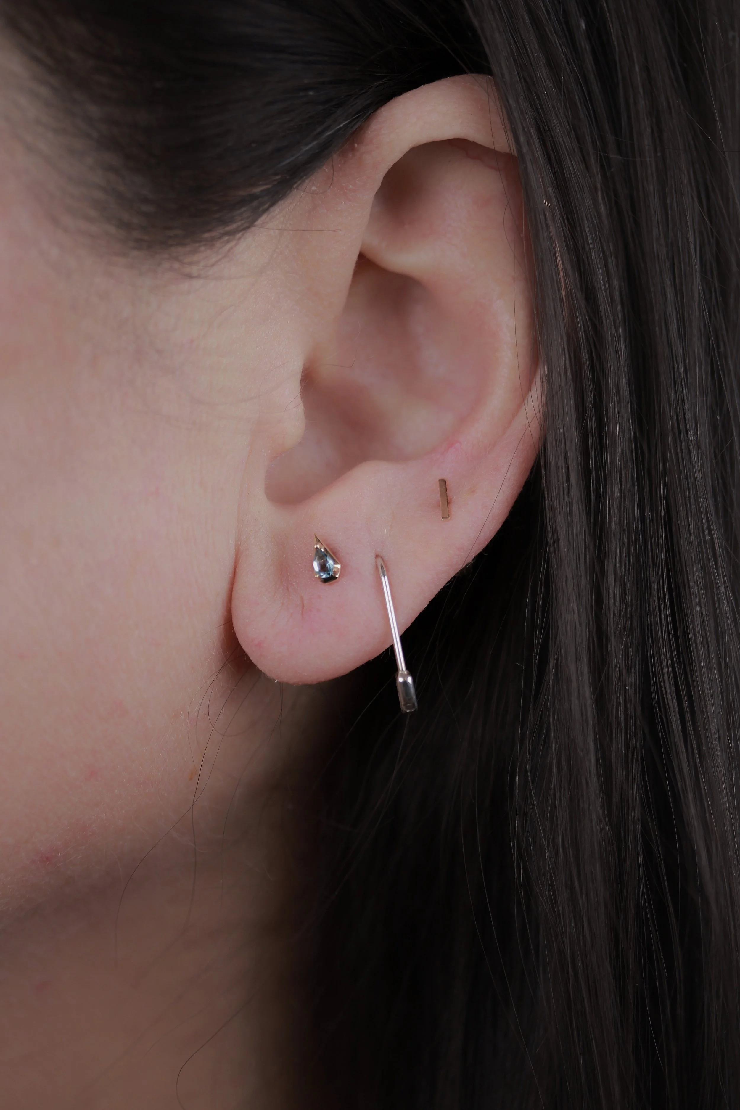 Close-up of a woman's ear with multiple earrings, including a teardrop-shaped gemstone, a thin bar, and a long, thin metallic dangle.