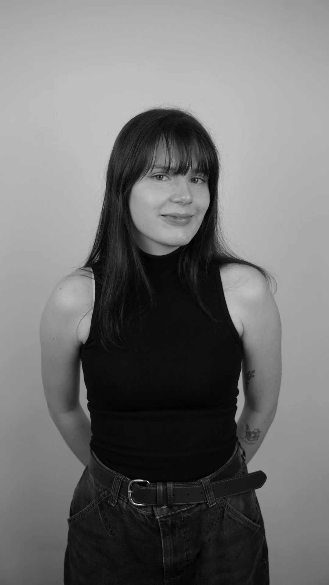 Black and white portrait of a woman with shoulder-length dark hair wearing a sleeveless black top and high-waisted pants with a belt, standing against a plain background.