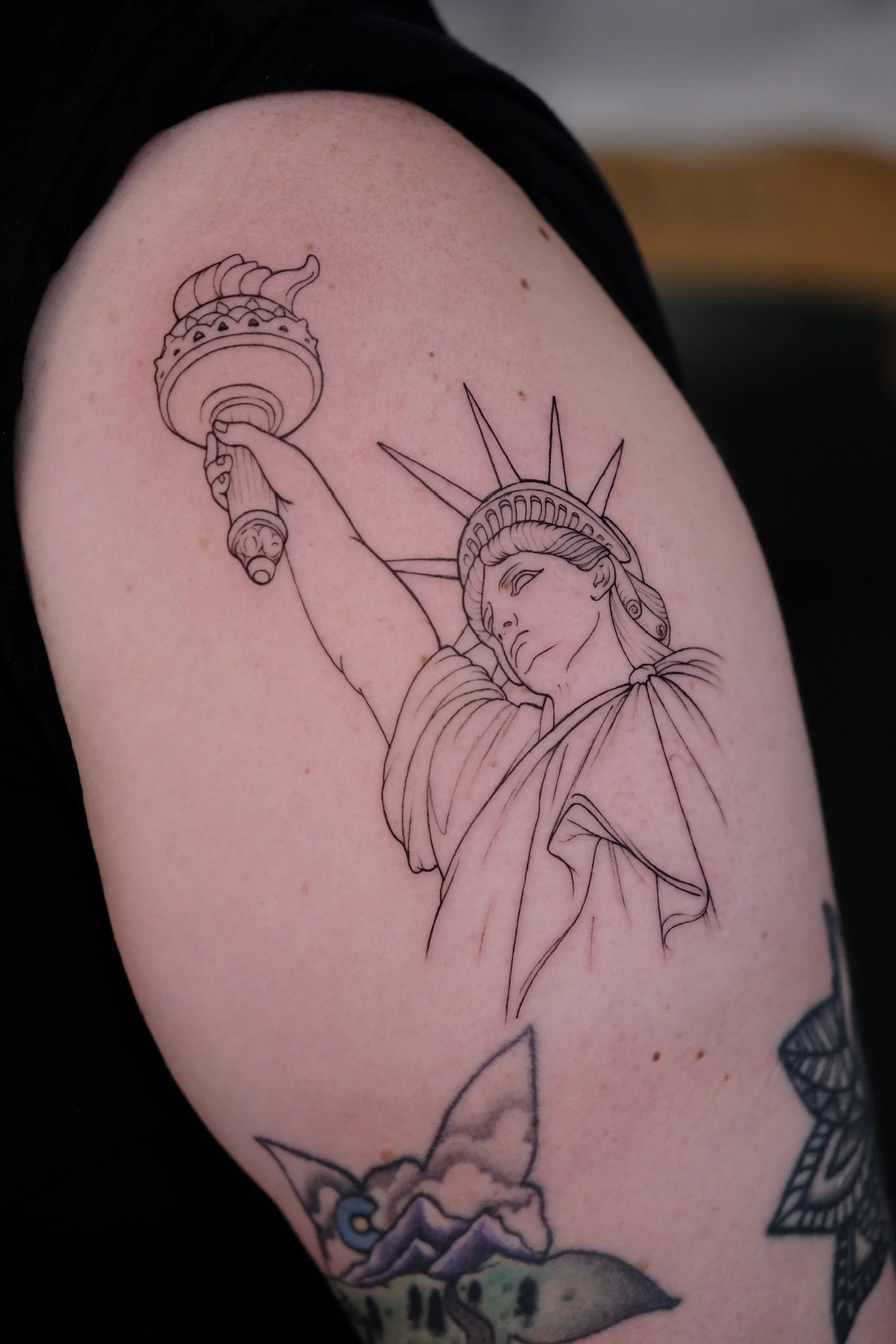 Line art tattoo of the Statue of Liberty holding a torch, with part of a traditional Japanese-style mountain scene beneath it.