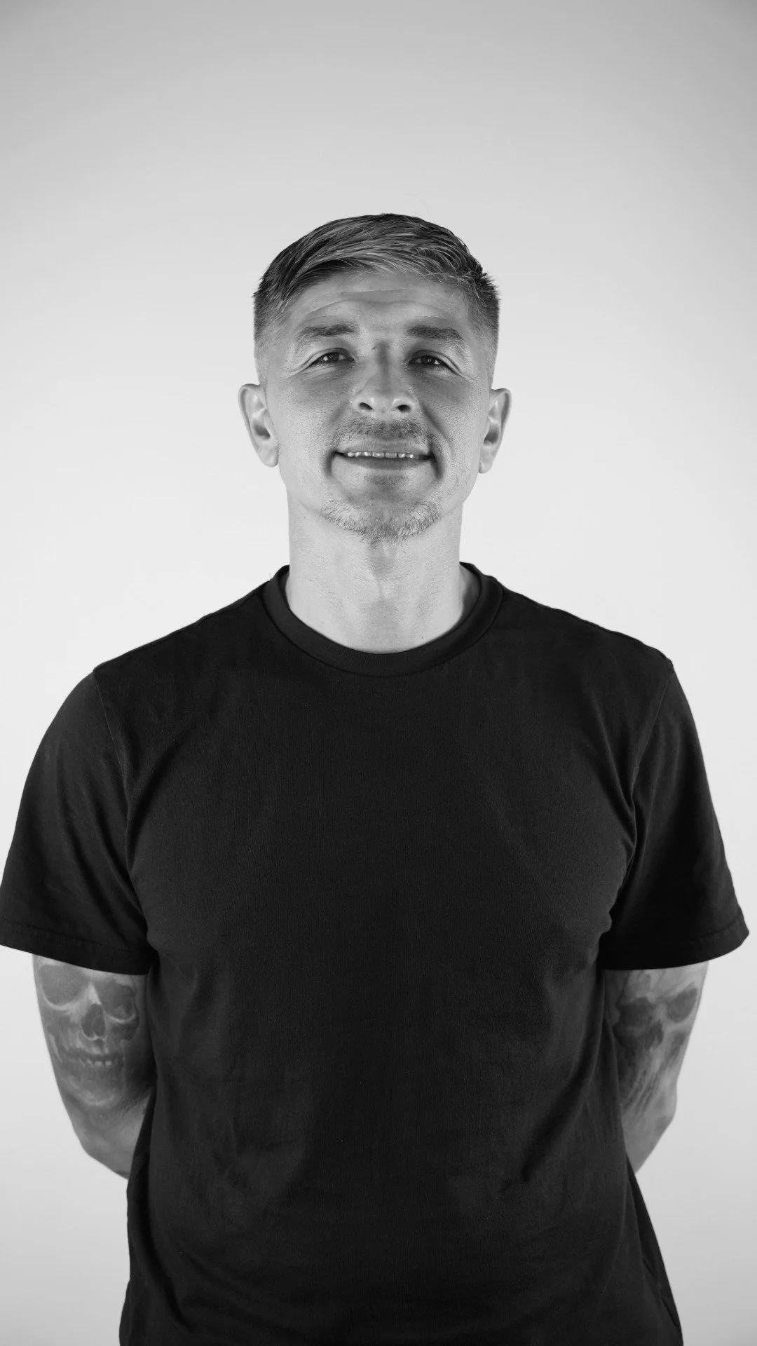 Black and white portrait of a man with short hair, light beard, wearing a black T-shirt, standing against a plain background.