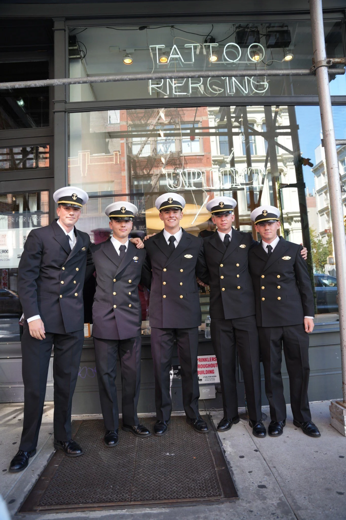 Five men in naval officer uniforms standing arm in arm outside a building with a neon sign that says 'TATTOO' and 'URGENCY' reflected in the glass window.