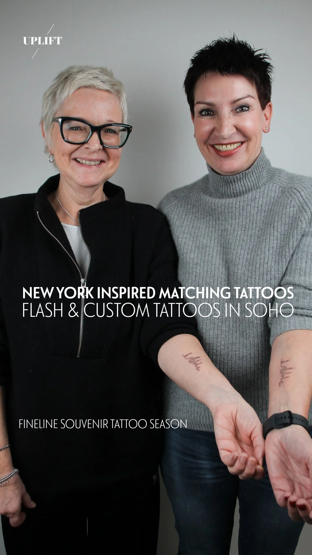 Two women display matching tattoos on their forearms, smiling at the camera against a plain background. One woman has short gray hair and glasses, wearing a black top, and the other has short dark hair, wearing a gray turtleneck sweater.