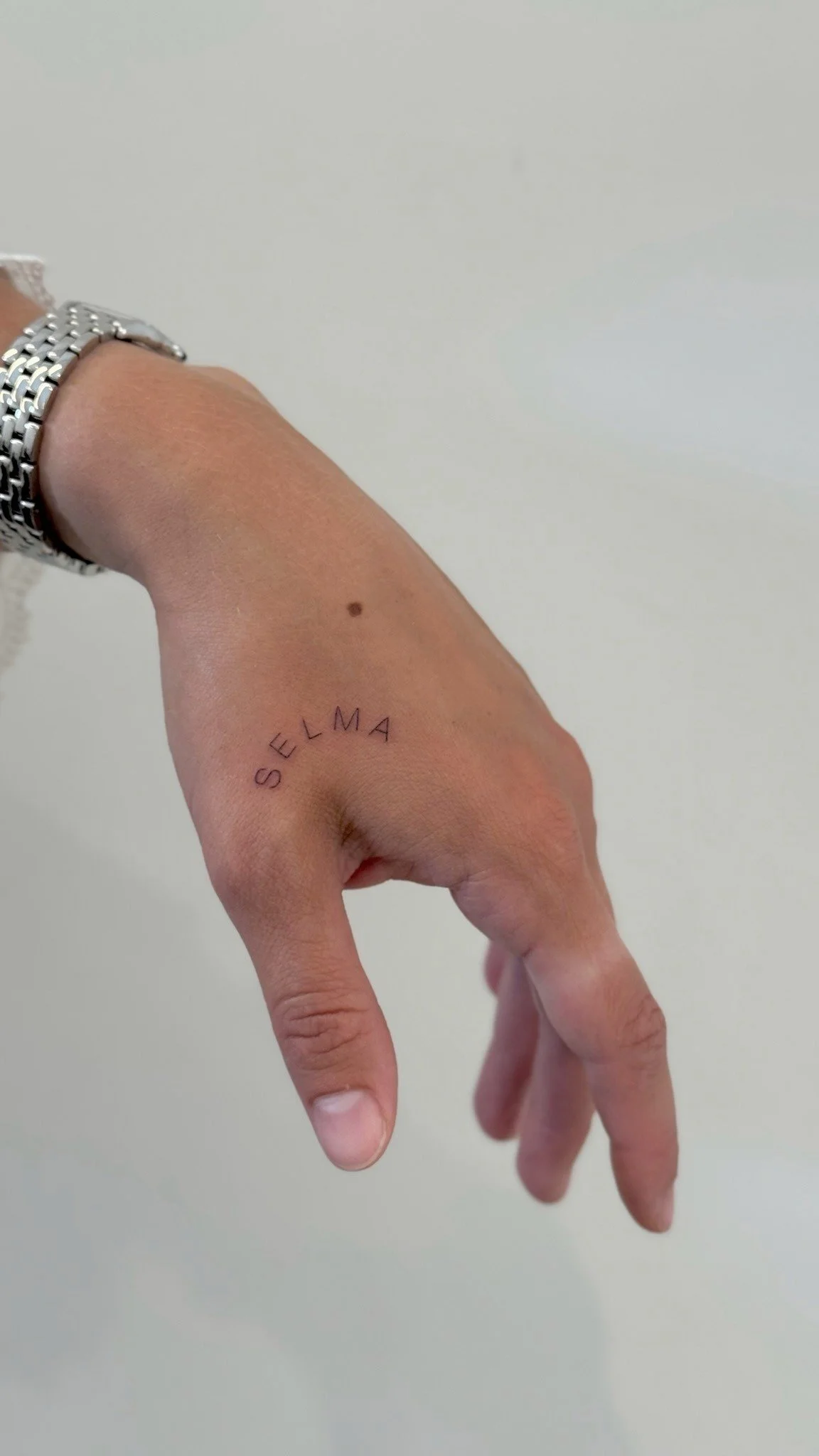 A person with a tattoo on their right hand that says 'SELMA' in capital letters. The hand is slightly bent with fingers in a relaxed position. The person is wearing a silver watch and has a mole on the wrist. The background is a plain light-colored w