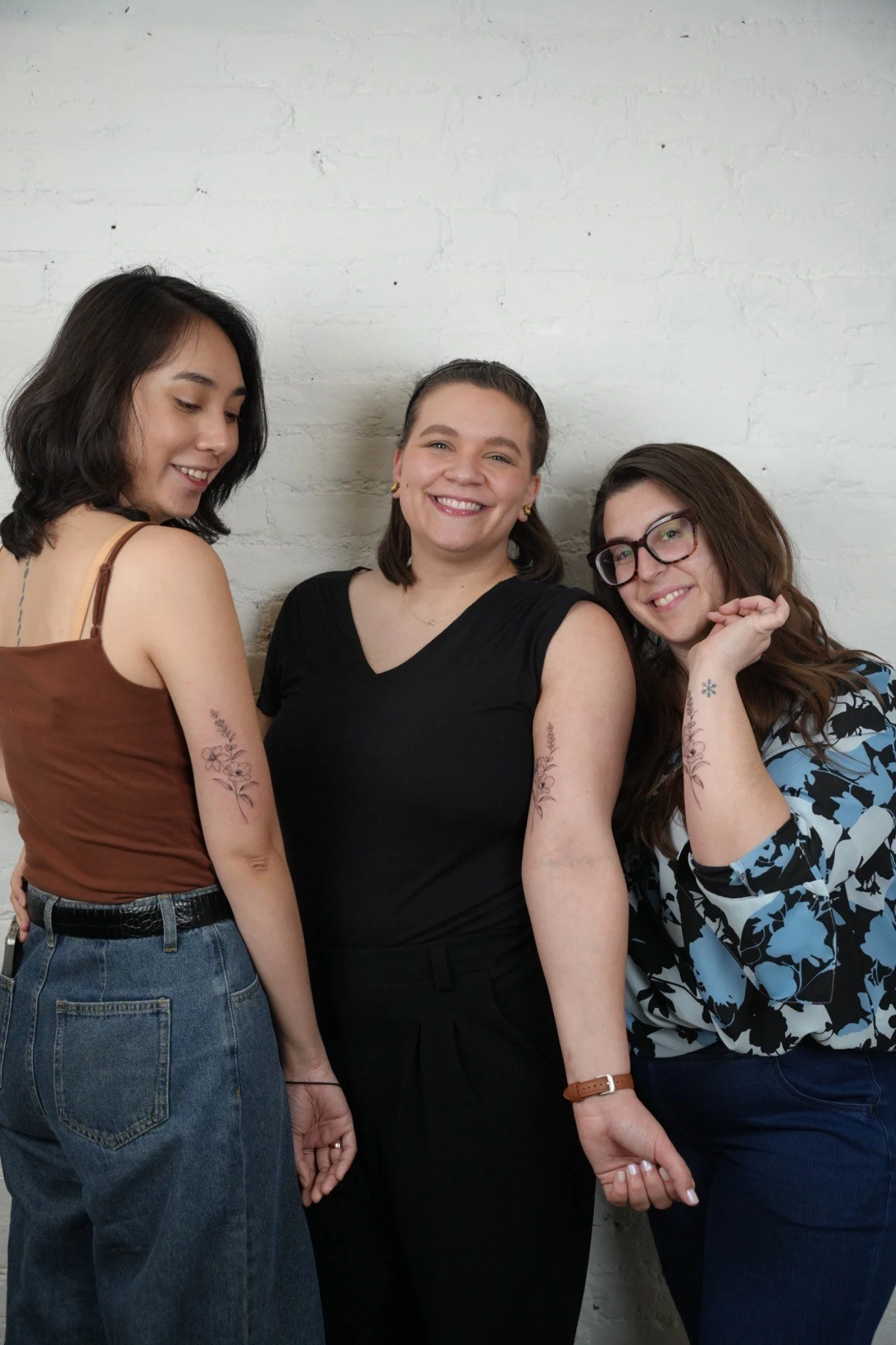 Three women smiling and standing close together against a white brick wall, with tattoos on their arms.
