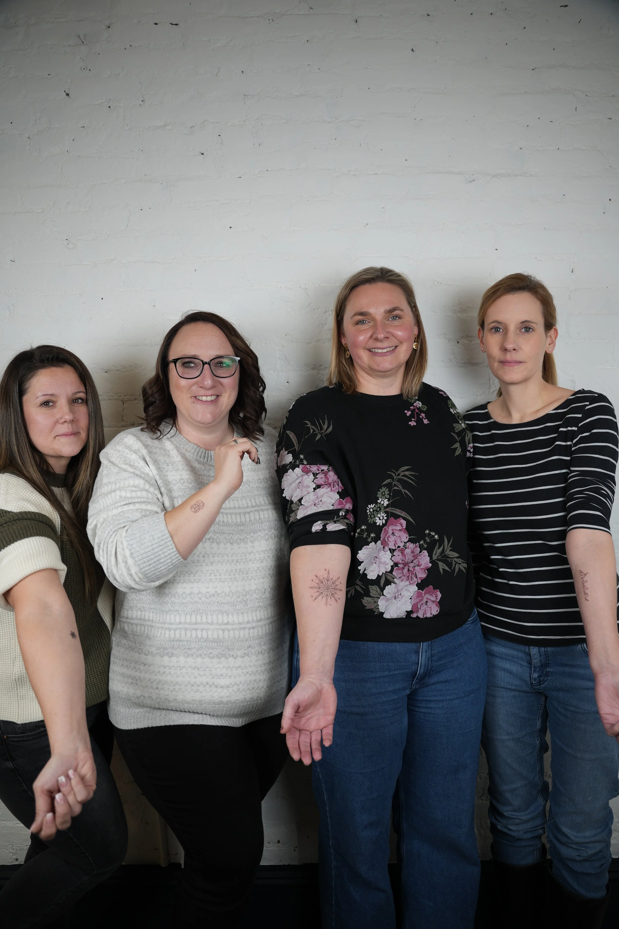 Four women standing against a white brick wall, posing for a group photo, dressed casually.