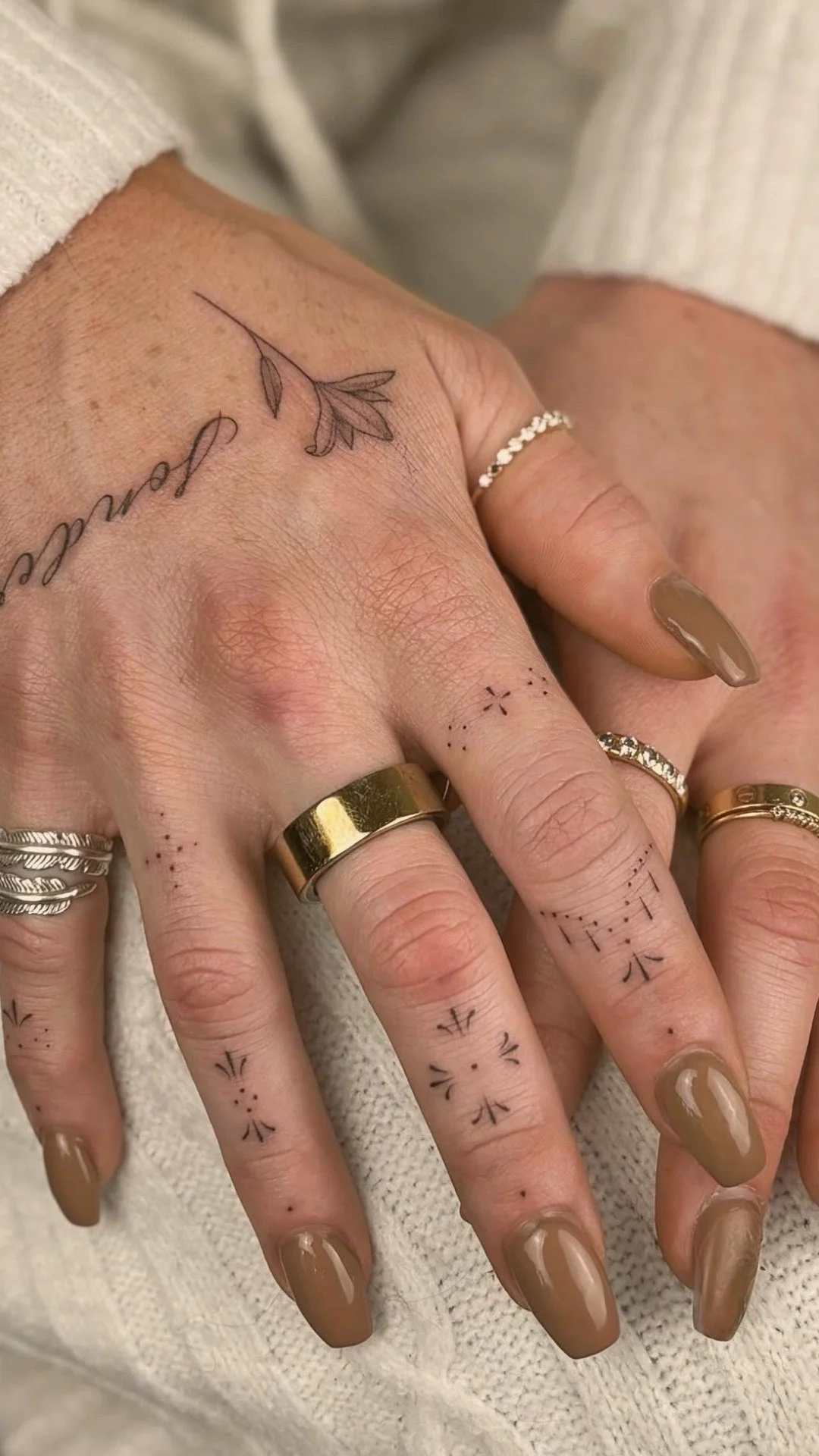A person's hand with several rings and tattoos, including a delicate leaf tattoo on the back of the hand and small geometric tattoos on the fingers, holding a beige textured fabric.