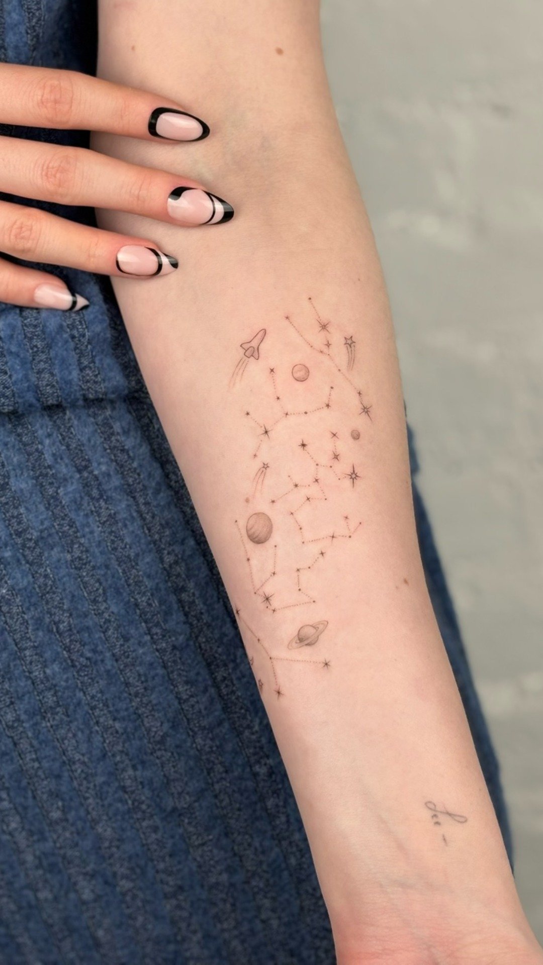 A person's forearm with a temporary tattoo of a constellation map featuring stars, planets, a rocket, and an alien eye, with a person wearing dark nail polish.