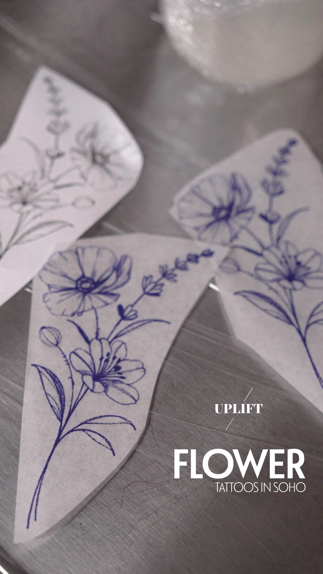 Temporary tattoo sheets with hand-drawn flower designs in purple, labeled as 'Uplift Flower Tattoos in Soho' on a metallic surface.