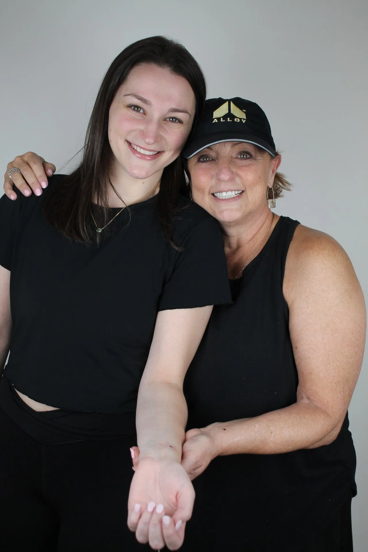 Two smiling women standing close together, one with her arm around the other. The older woman is wearing a black cap with gold lettering and a sleeveless black top, while the younger woman has shoulder-length dark hair and is wearing a black shirt. They are against a plain light gray background.
