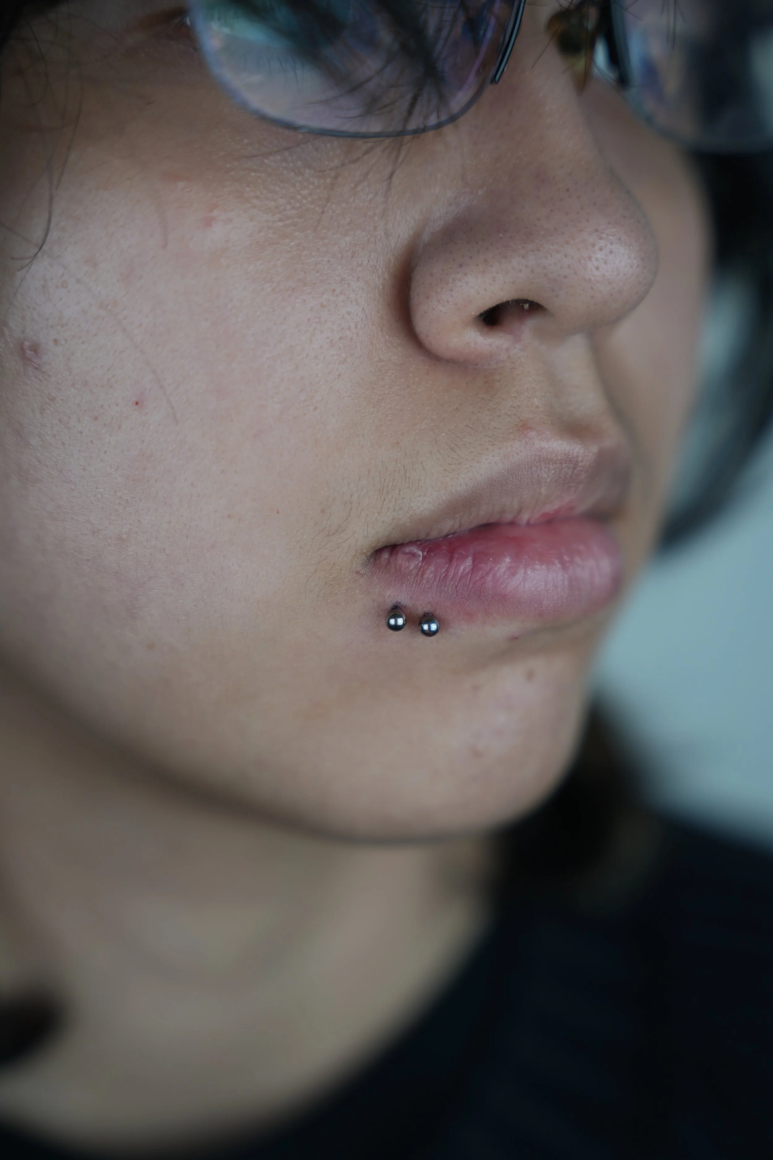 Close-up of a person's face showing their nose, lips, and a double lip piercing with two metal balls, with glasses partially visible at the top.