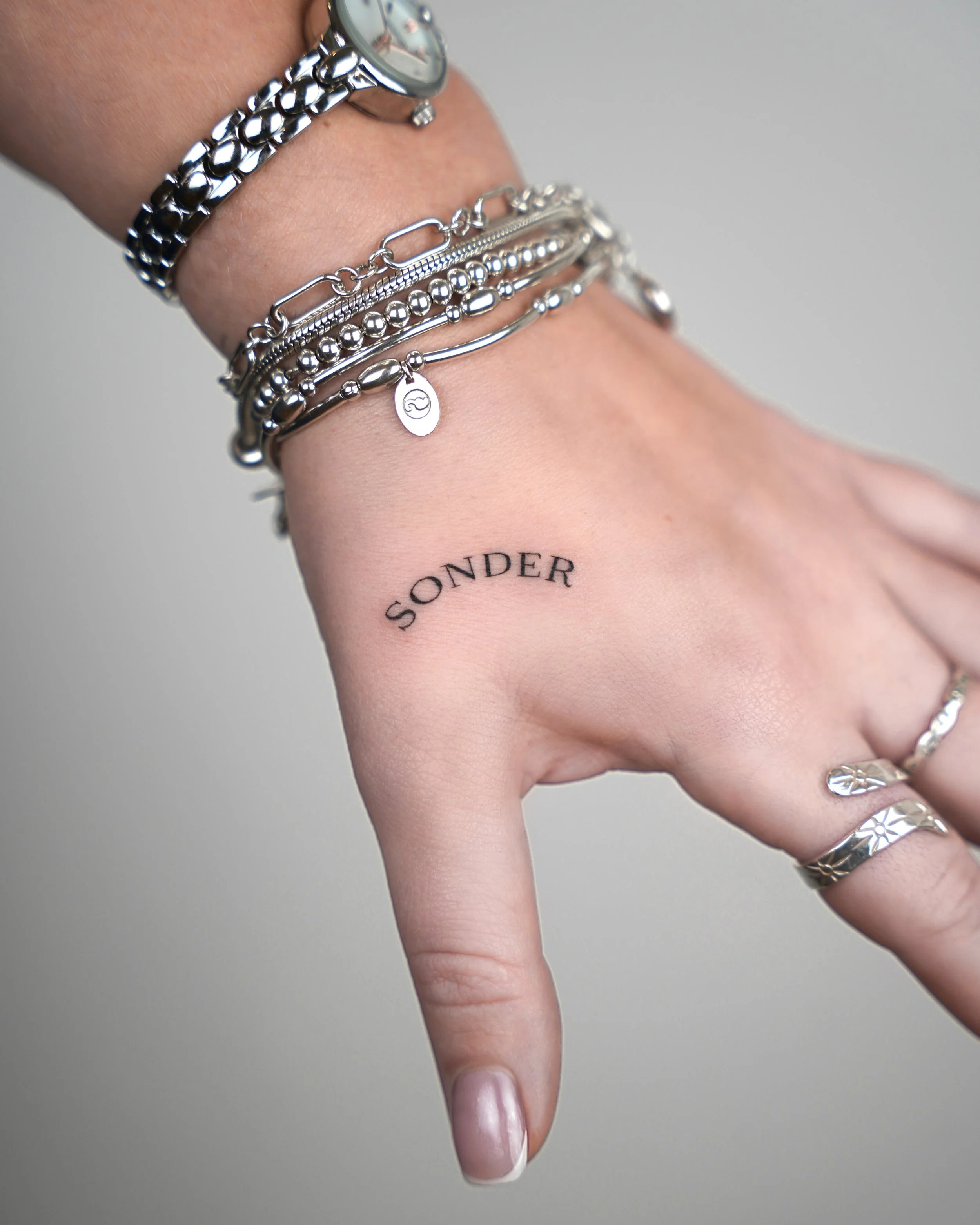 A person's hand and wrist with multiple silver bracelets and rings, displaying a temporary tattoo with the word 'SONDER' on the wrist.