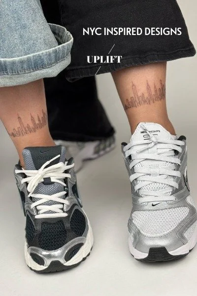 Person wearing sneakers and rolled-up jeans, showing NYC-inspired tattoos on ankles, with black shorts featuring text about NYC-inspired designs and Uplift.