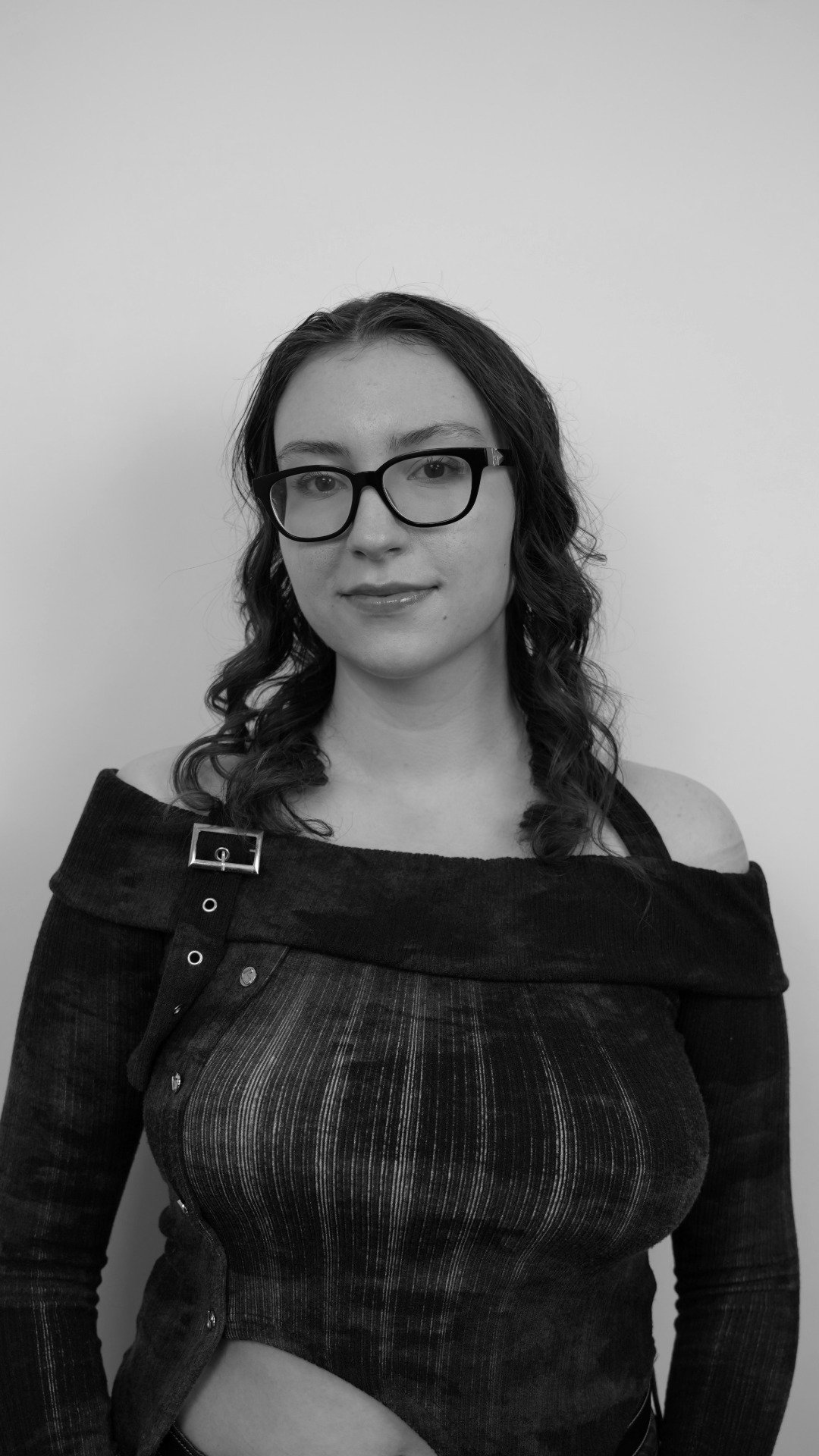 Black and white portrait of a woman with dark hair and glasses, wearing a dark off-shoulder top with buttons and a pin, standing against a plain light-colored background.