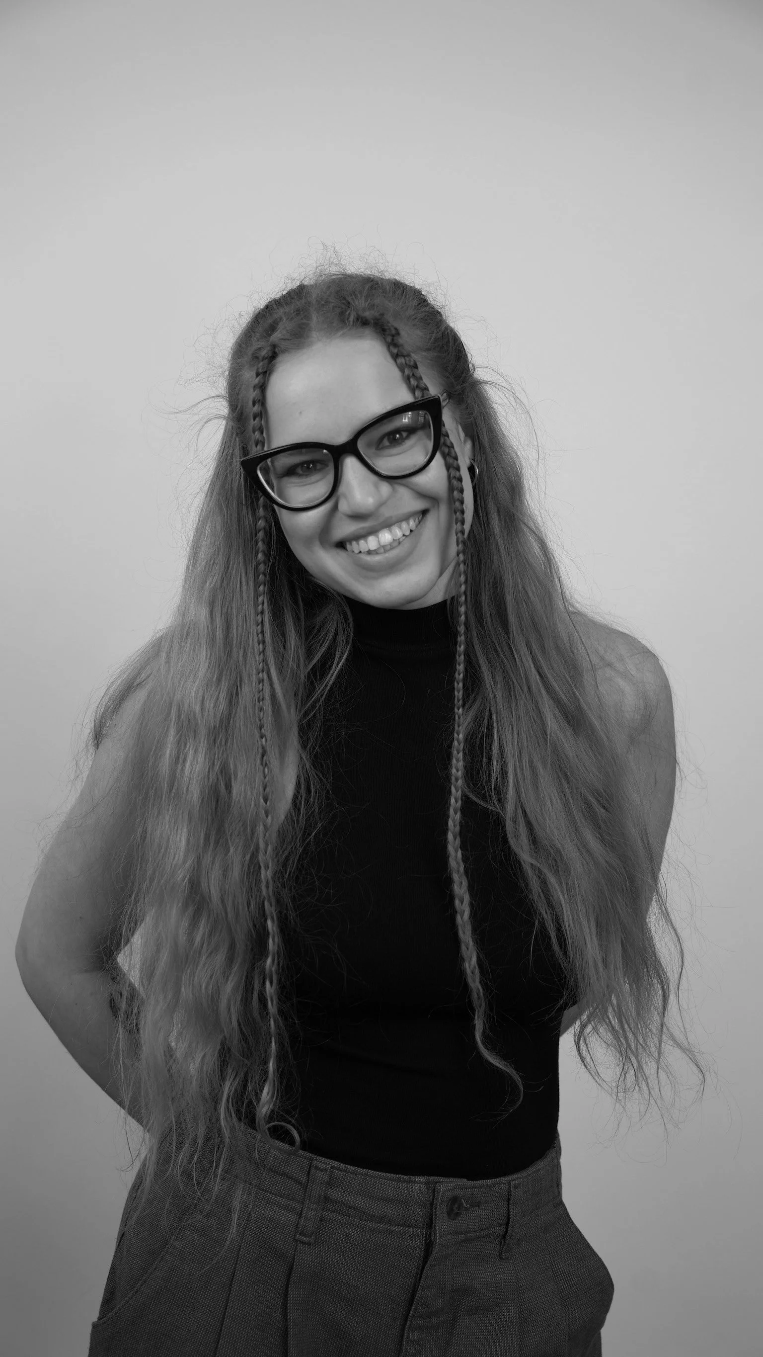 Young woman with curly hair and braided strands wearing glasses, smiling, dressed in a sleeveless top and checked trousers, posed against a plain background.