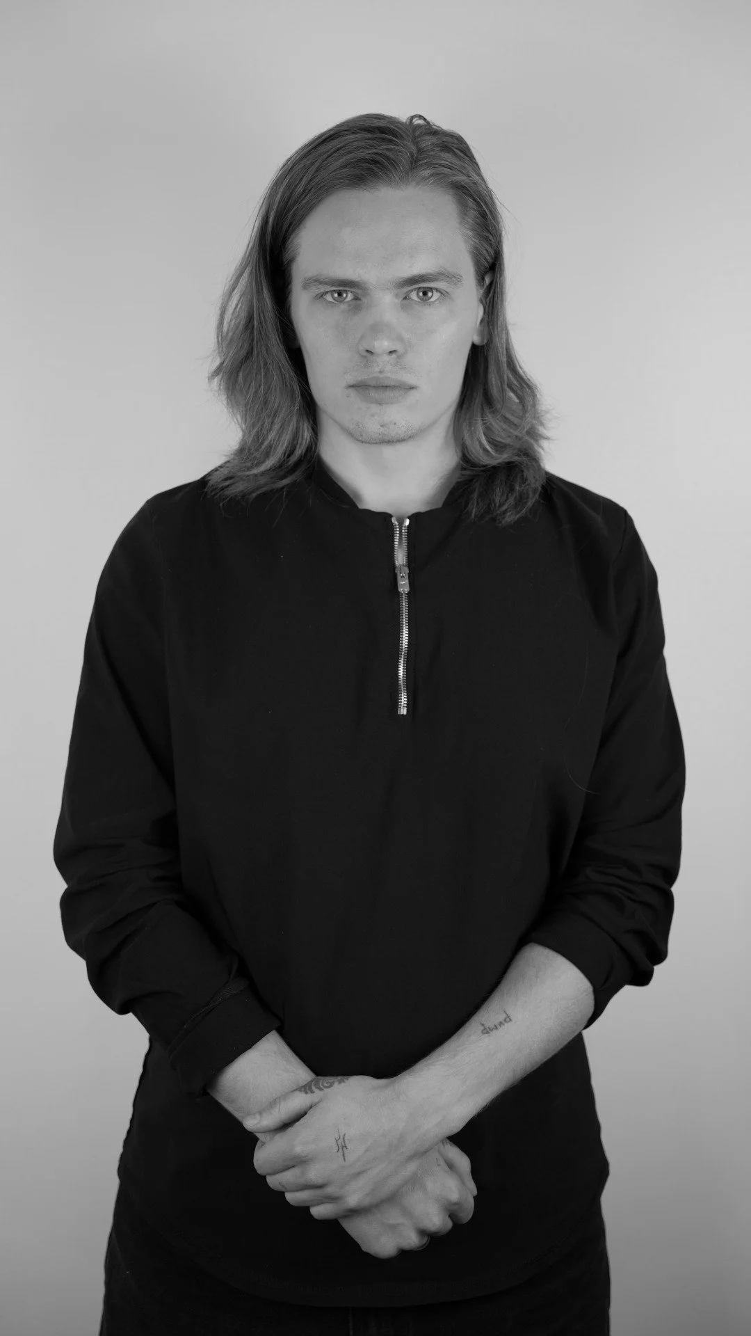 Black and white portrait of a young man with long hair, wearing a zippered black jacket, standing against a plain light background.