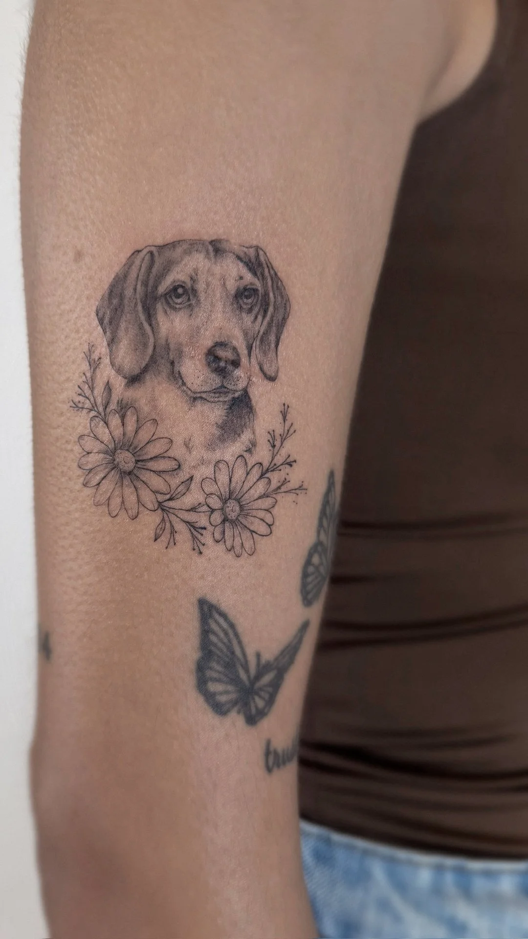 Tattoo of a realistic dog portrait surrounded by flowers on person's upper arm, with additional butterflies tattooed below.