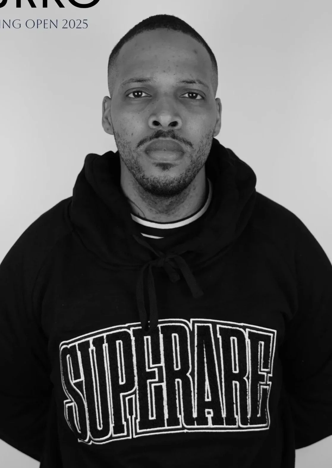 @turkotattoonyc Turko Resident Tattoo Artist Black and white portrait of a man with short hair and facial hair, wearing a hoodie with 'SUPREME' logo and striped t-shirt underneath, against a plain background.