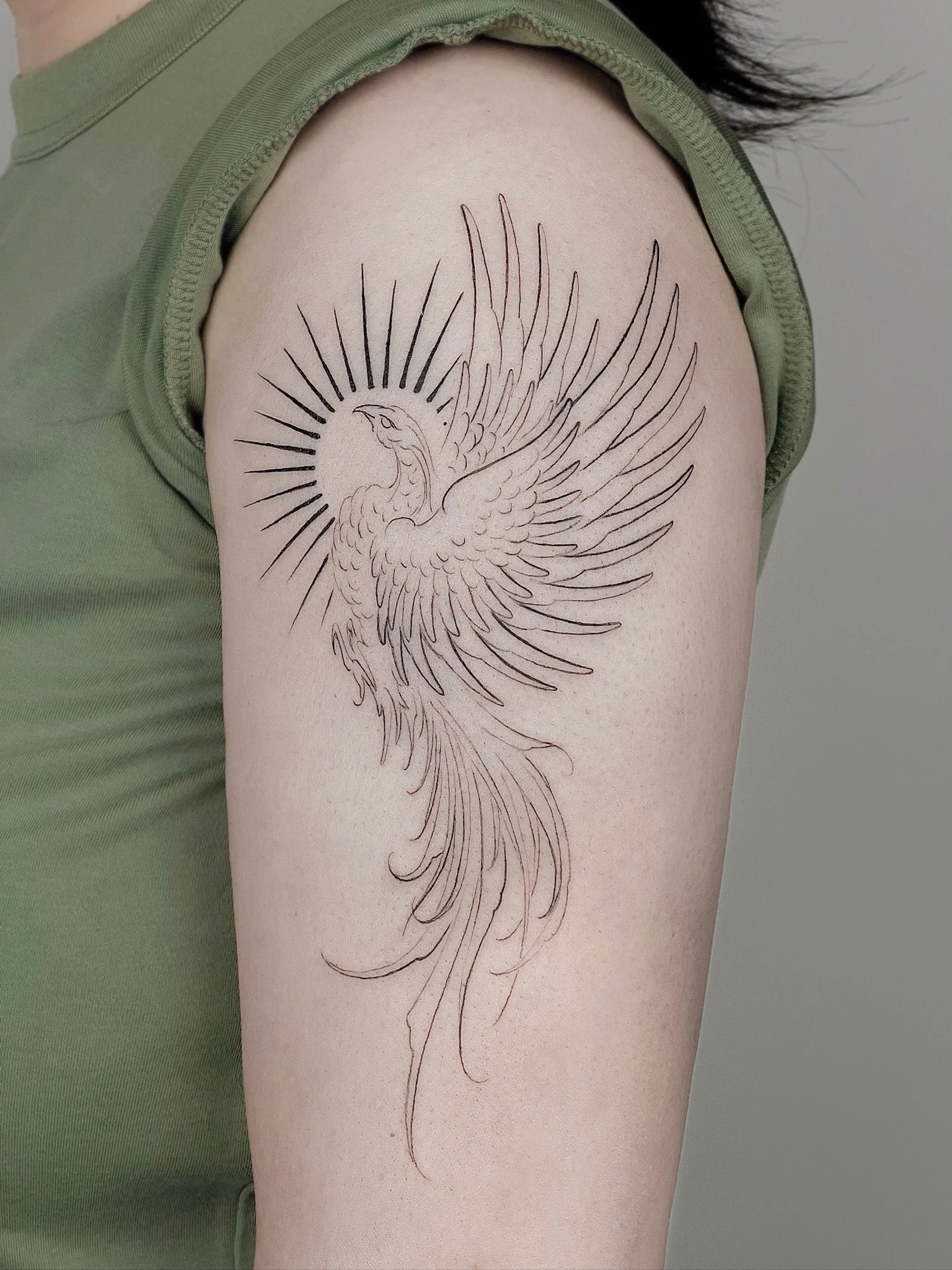 Tattoo of a phoenix with its wings spread, surrounded by radiant lines, on a person's upper arm.