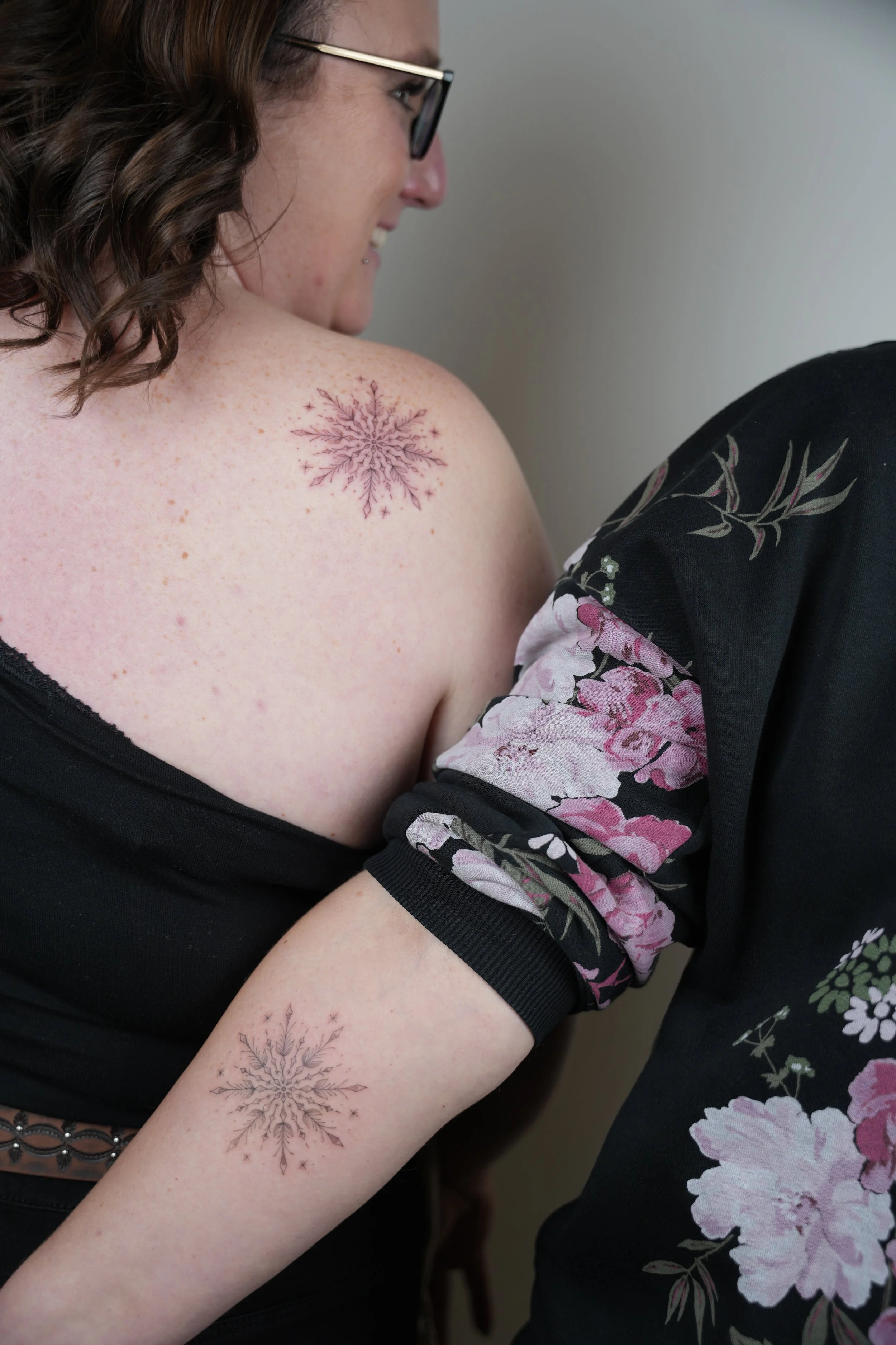 A woman with glasses and brown curly hair shows a snowflake tattoo on her shoulder and an identical snowflake tattoo on her upper arm, while smiling and leaning towards someone wearing a black floral shirt.