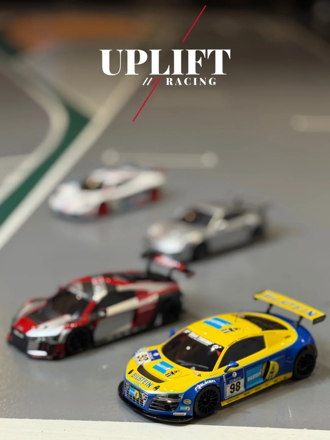 Model race cars on a miniature race track, with a yellow and blue car in the foreground and other colored cars in the background, branded with Uplift Racing logo.