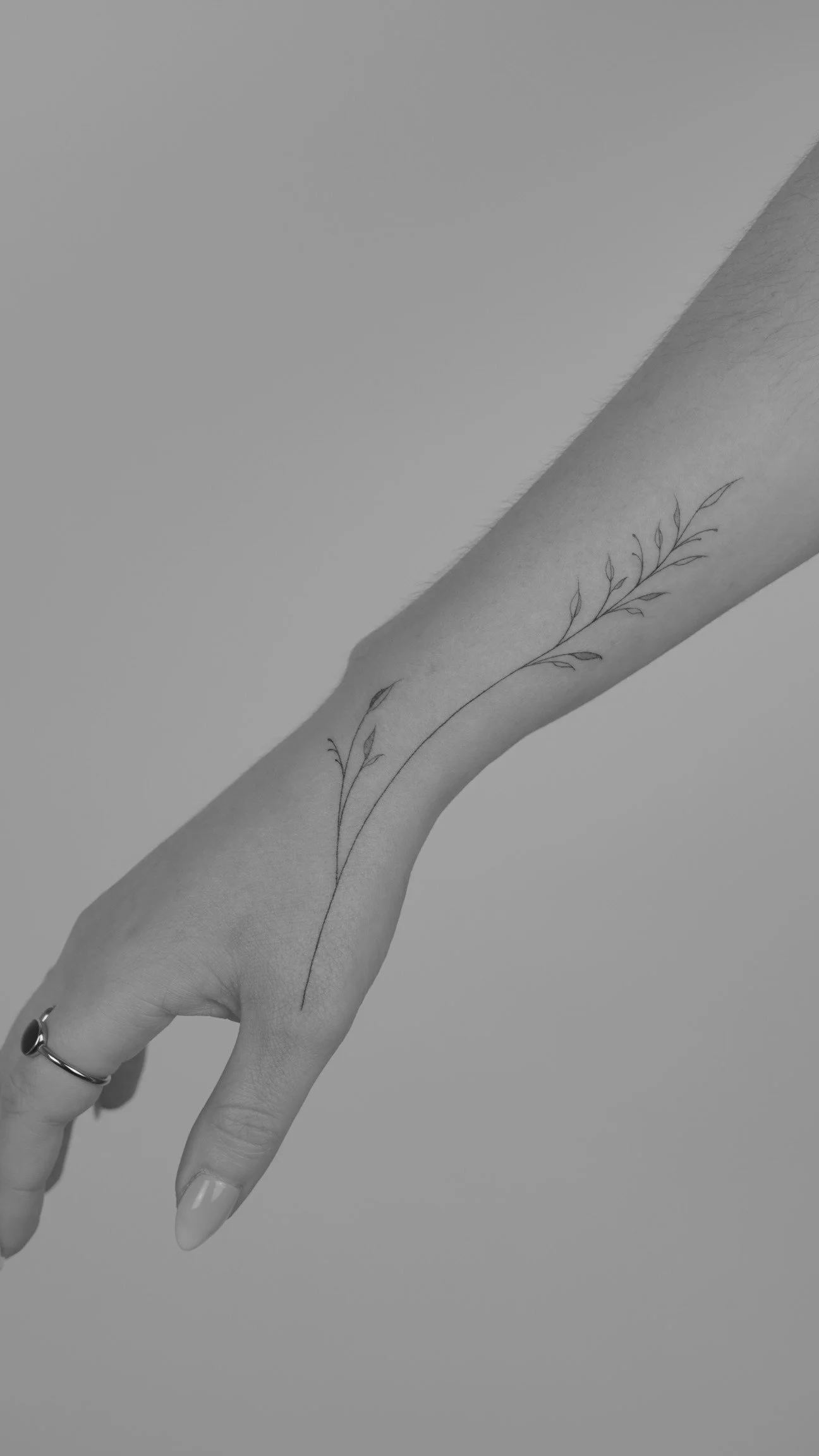 Black and white photograph of a person's arm with a delicate, minimalist floral tattoo running along the forearm.