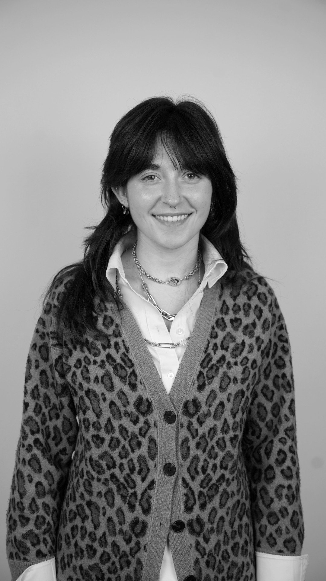 Black and white portrait of a woman smiling with dark, shoulder-length hair, wearing a leopard print cardigan over a white shirt, layered with multiple necklaces and earrings.