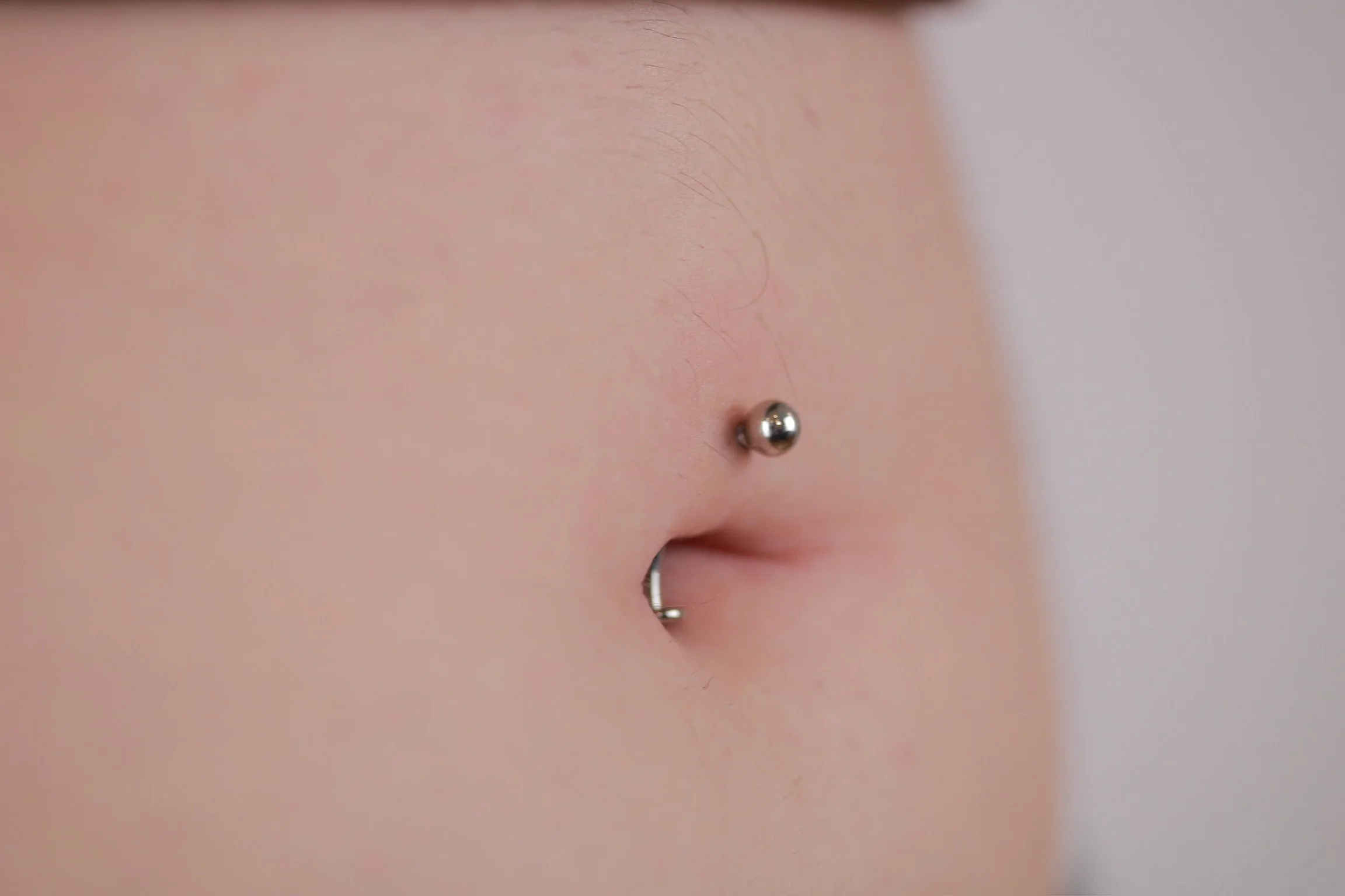 Close-up of a person's midsection showing a navel piercing with a silver hoop and a small nose piercing with a silver ball.