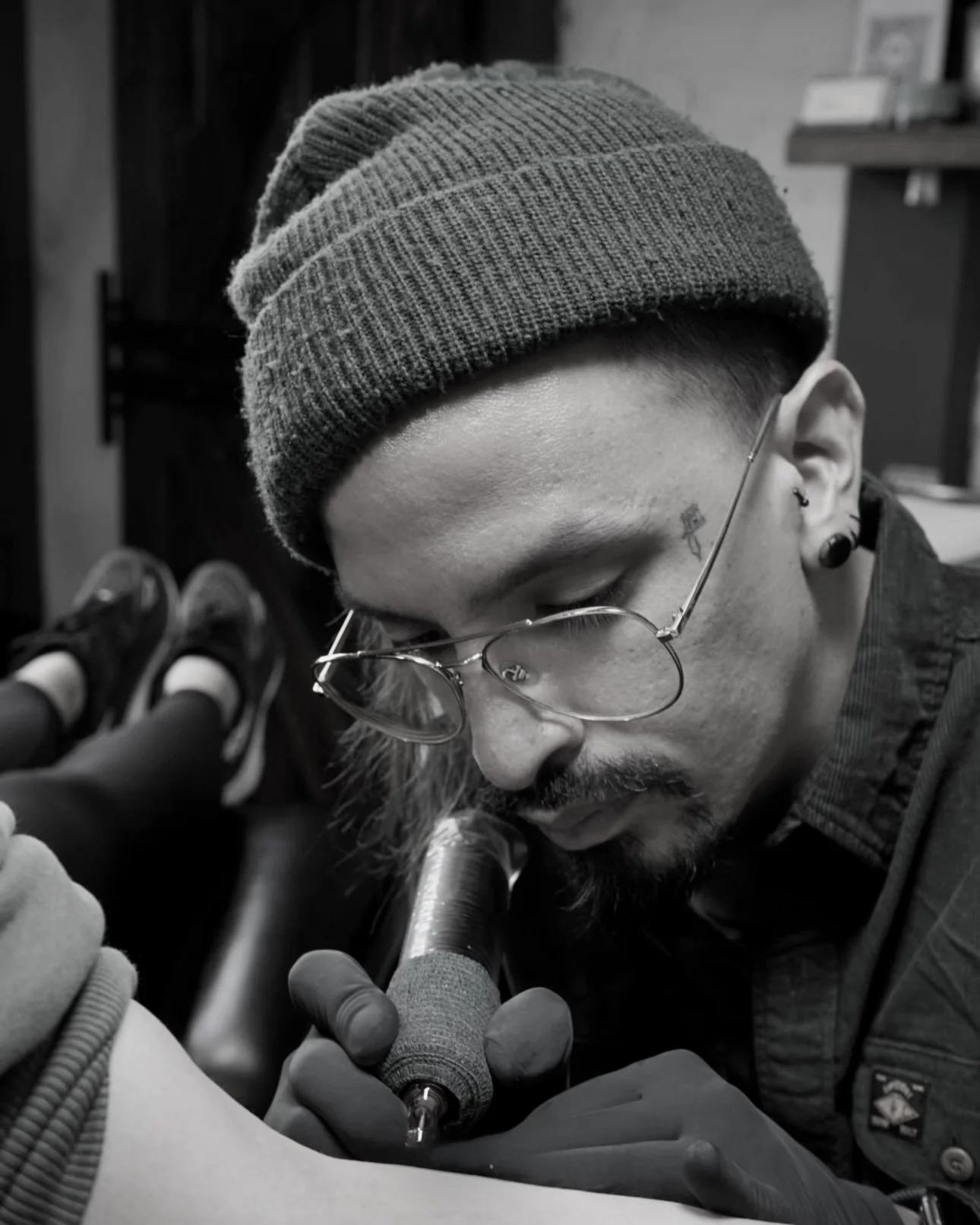 Christopher - Resident Tattoo Artist A person with glasses, a small tattoo near the eye, and ear piercings gets a tattoo on their arm in a tattoo studio. The person wears a beanie and gloves and leans forward with their eyes closed.