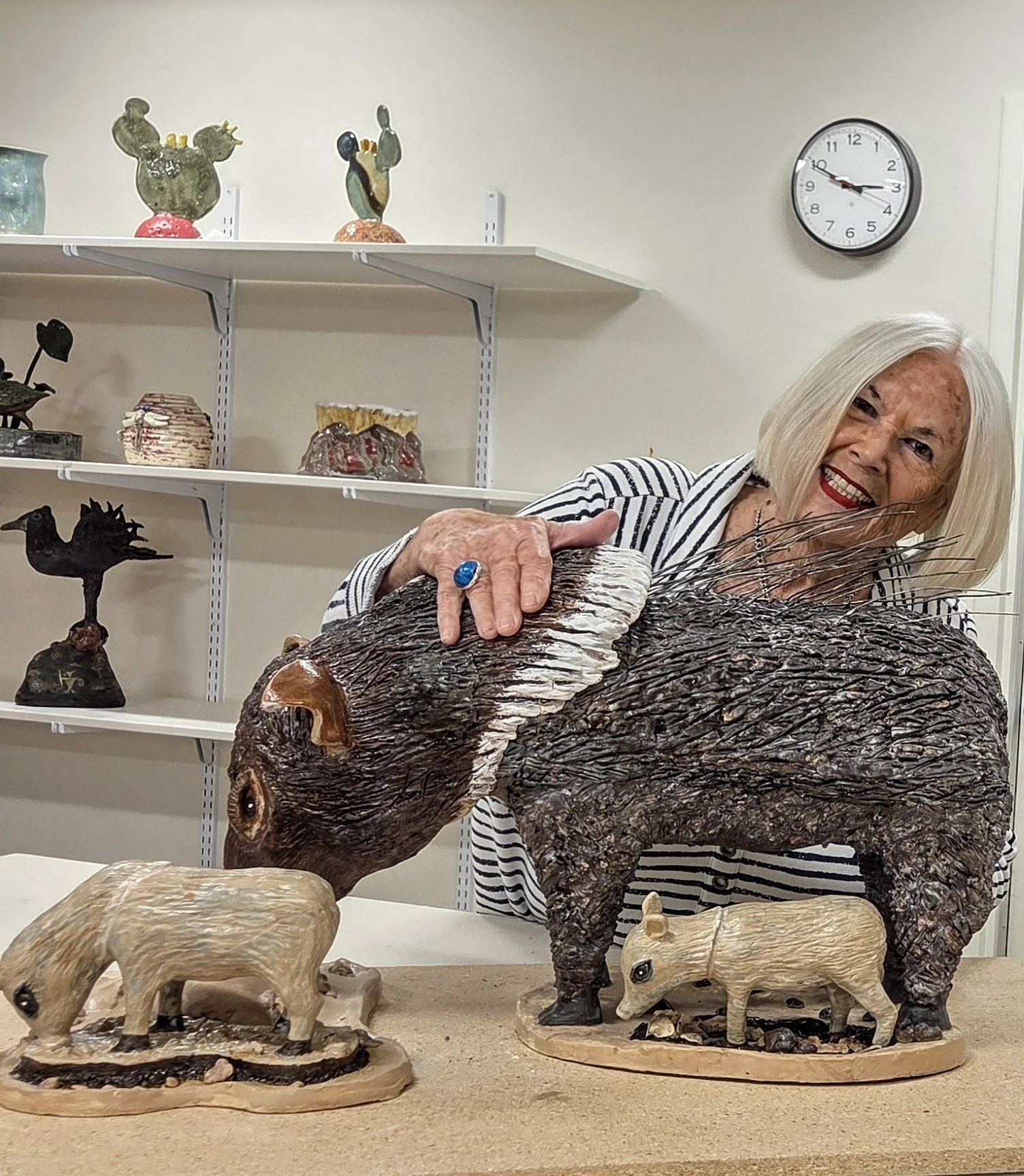 An elderly woman smiling and holding a large wooden sculpture of a lion. On the table, there are several smaller wooden animal sculptures, including a lion and a boar. In the background, there are decorative pottery and sculptures on white shelves, and a wall clock showing 3:49.