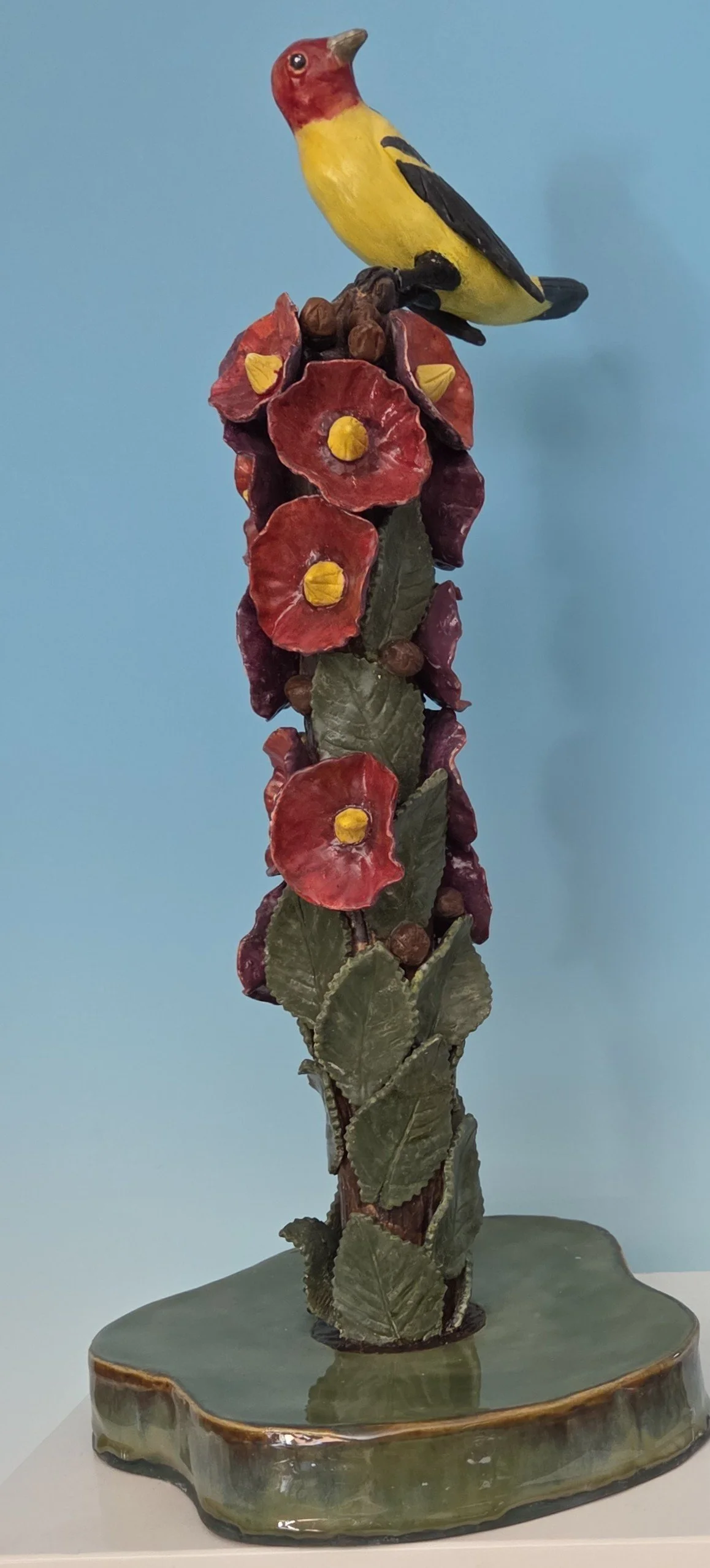 A decorative sculpture of a yellow bird with a red head and black wings, perched on a branch among red flowers with yellow centers, mounted on a green base.