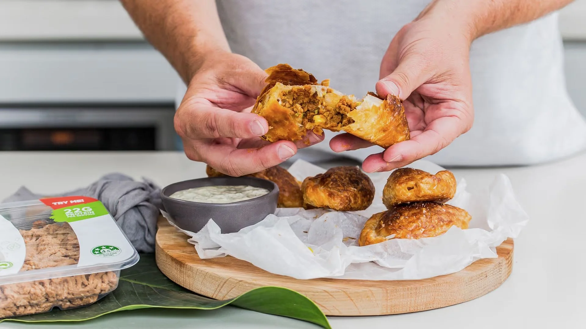 A person holding a piece of savory meat pie with a flaky crust, revealing a filling of ground meat, corn, and vegetables, with additional meat pies and a side of dipping sauce on a wooden platter.