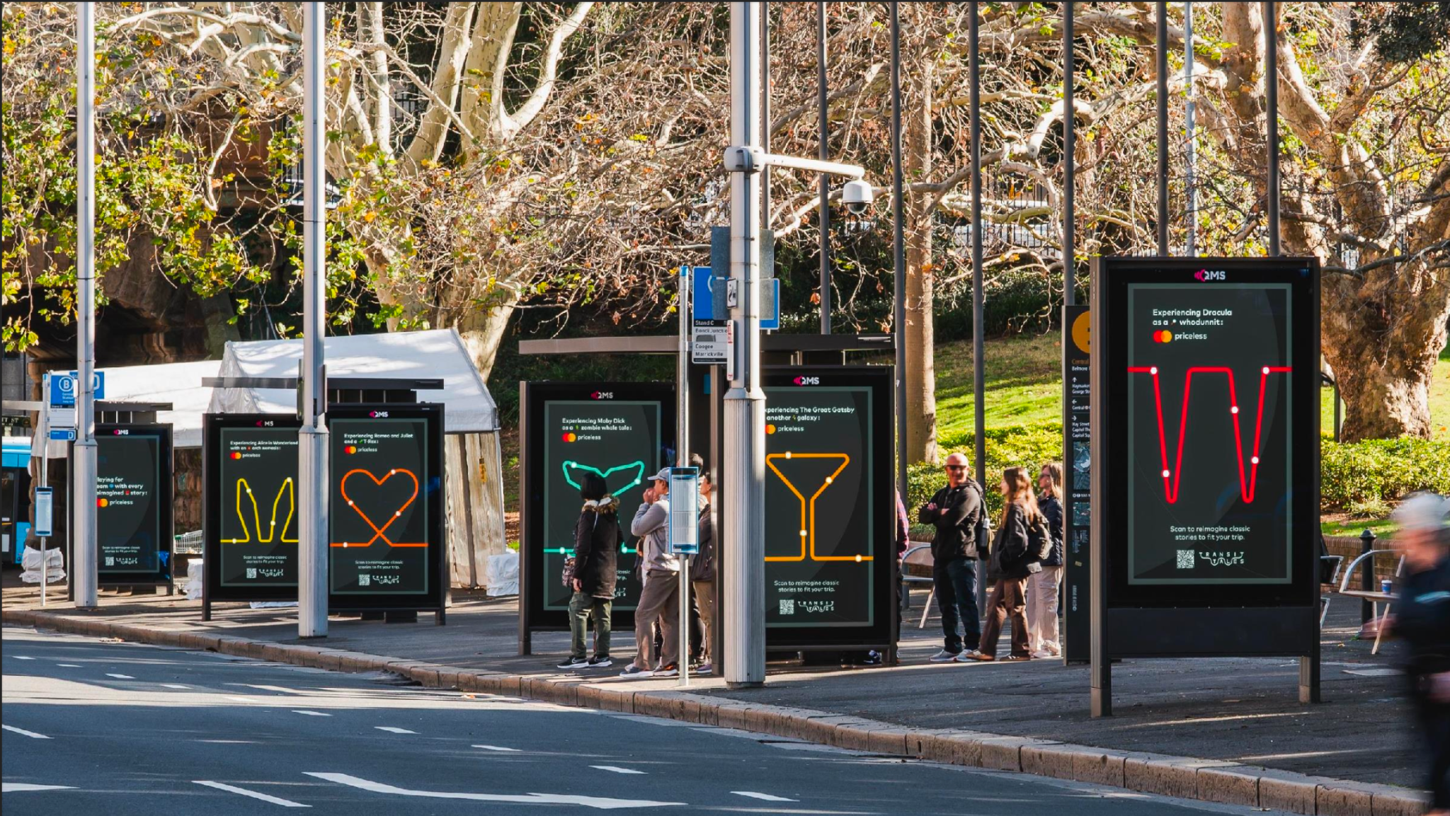 Bus stop with digital advertising displays on a city sidewalk, featuring classic story themes with vibrant neon-style illustrations, and people waiting or walking nearby in an outdoor urban setting with trees.