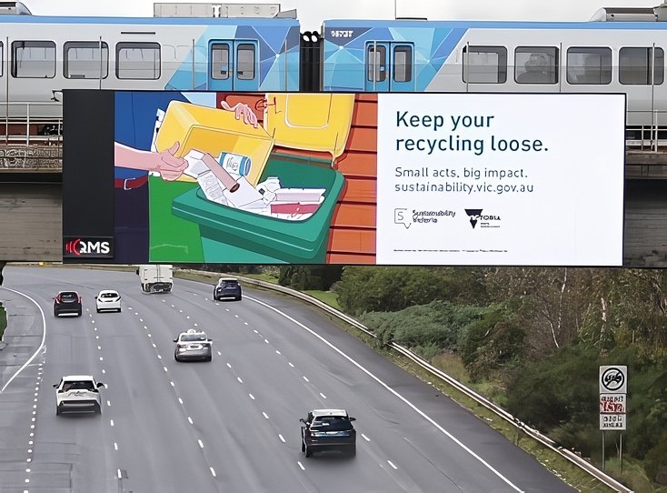 A highway with several cars and a large billboard that says, "Keep your recycling loose" and features an illustration of a hand placing paper into a recycling bin.