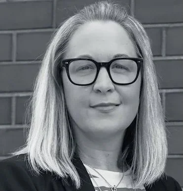 HERO Creative Director Sue Young, a woman with light-colored, shoulder-length hair wearing glasses and a dark top, standing in front of a brick wall.