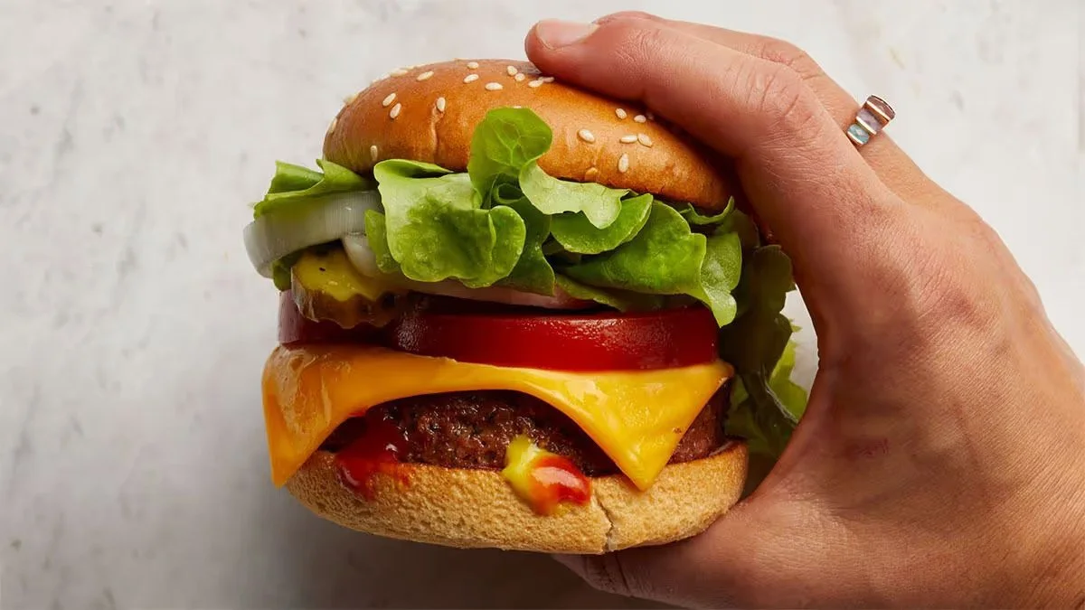 Hand holding a cheeseburger with lettuce, pickles, onion, tomato, cheddar cheese, beef patty, and sandwich bun.