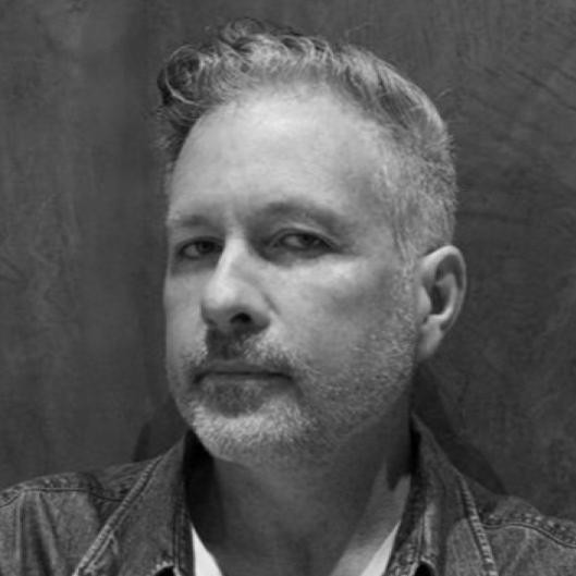 A black and white close-up portrait of HERO Creative Director Andrew Woodhead with short, styled hair, slight facial hair, and wearing a denim jacket, looking confidently at the camera.