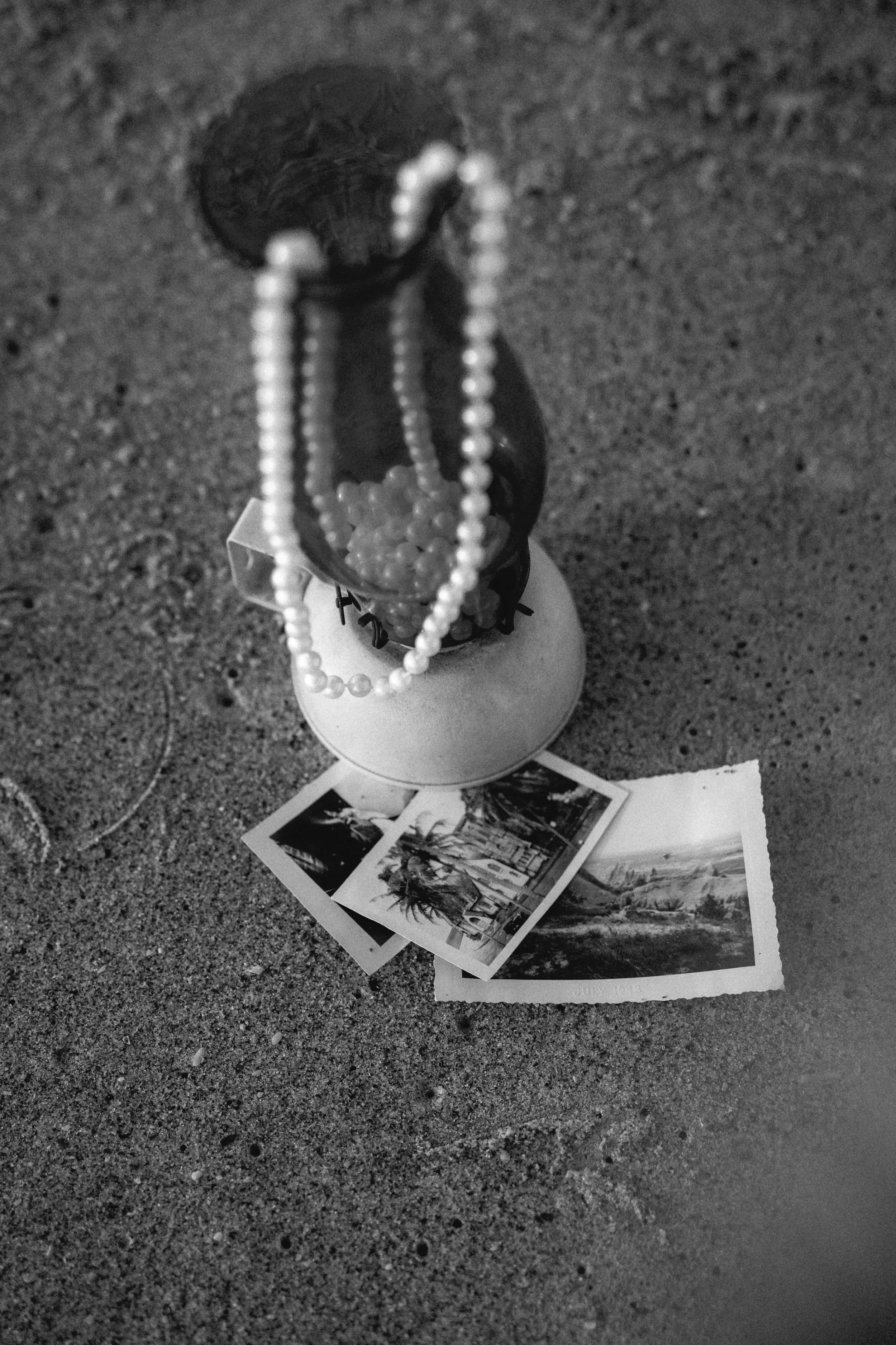 Black and white photo of a small display with photographs on the ground, a sculpture with pearl necklaces, and a small figure or object on a circular platform, all set on a sandy surface.