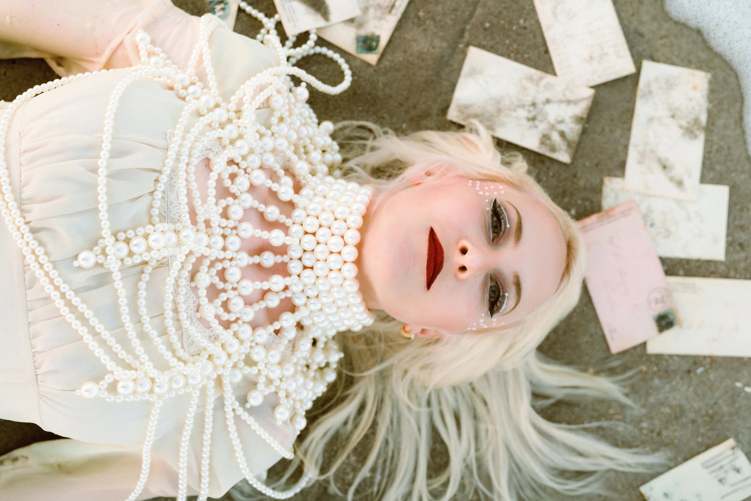 A woman dressed in a white outfit decorated with multiple strands of large white pearls lies on a surface, with her face and part of her upper body visible. She has platinum blonde hair, dark lipstick, and eye makeup with rhinestones around her eyes.