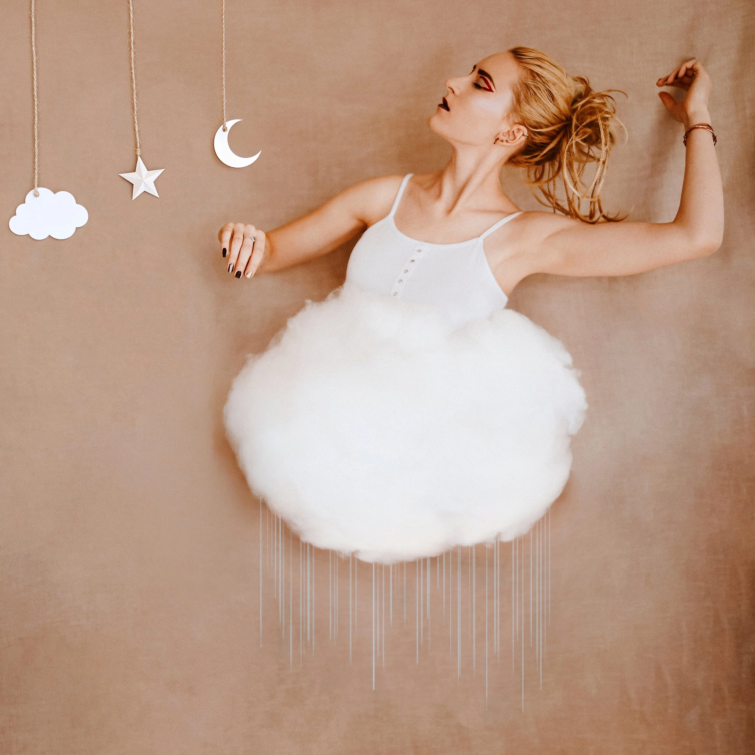 A woman with red hair and dramatic makeup lying on her side with her right arm raised above her head, wearing a white top, against a neutral background. Hanging from the ceiling are white paper cutouts of a cloud, a star, a moon, and a crescent moon.