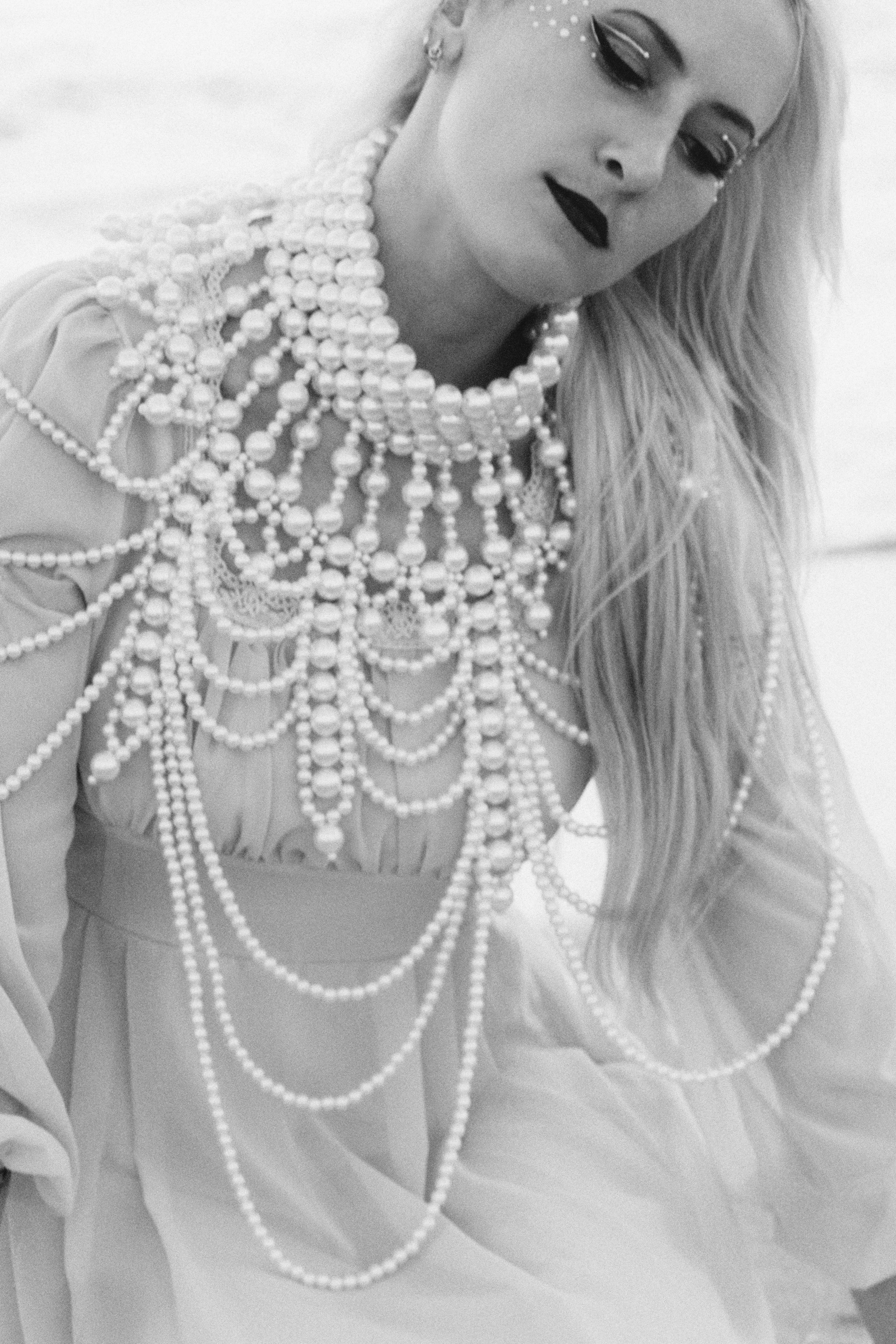 A young woman with long hair wearing multiple strands of pearl necklaces, dark lipstick, and eye makeup, smiling with eyes closed.