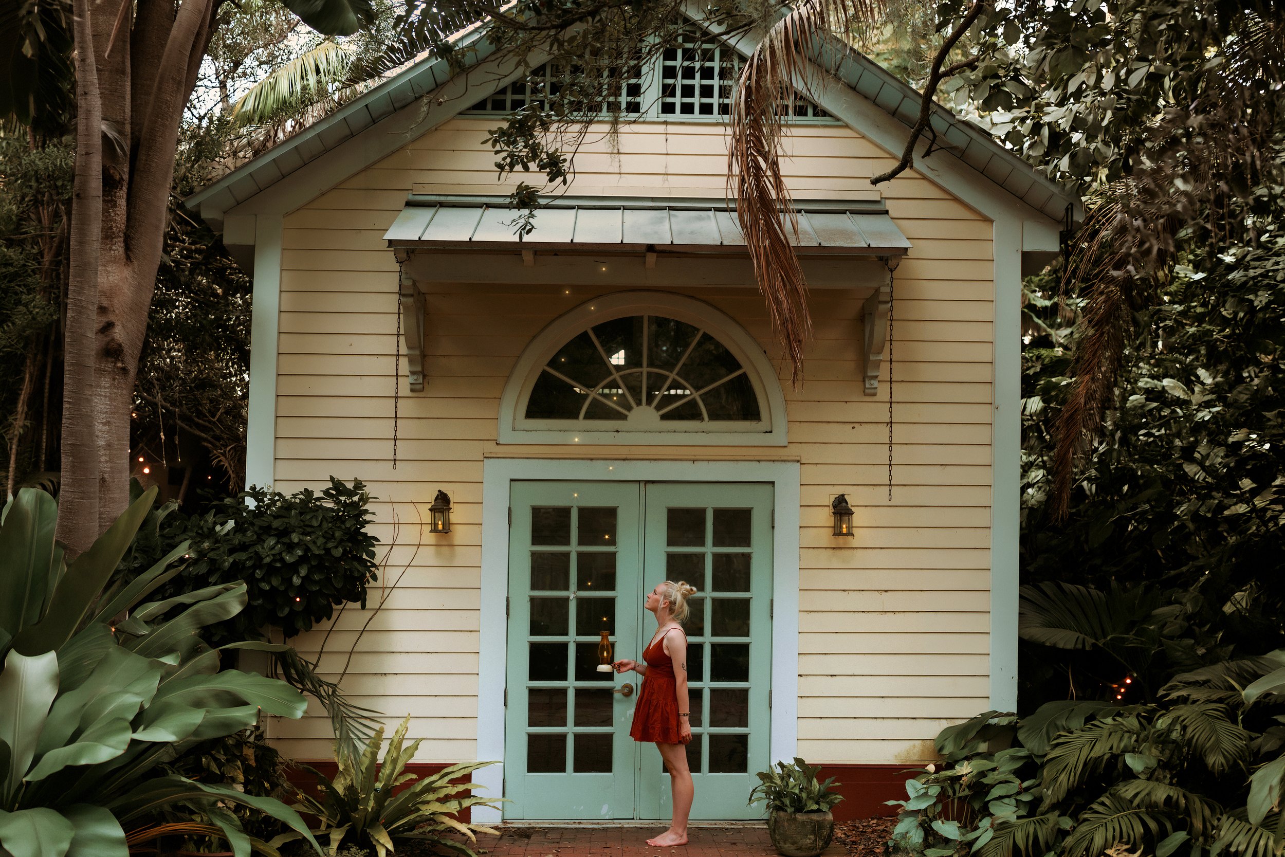 A woman in a rust-colored dress holding a tray with a bottle and a glass, standing in front of a pastel green door and window in a yellow house surrounded by lush green plants and trees.