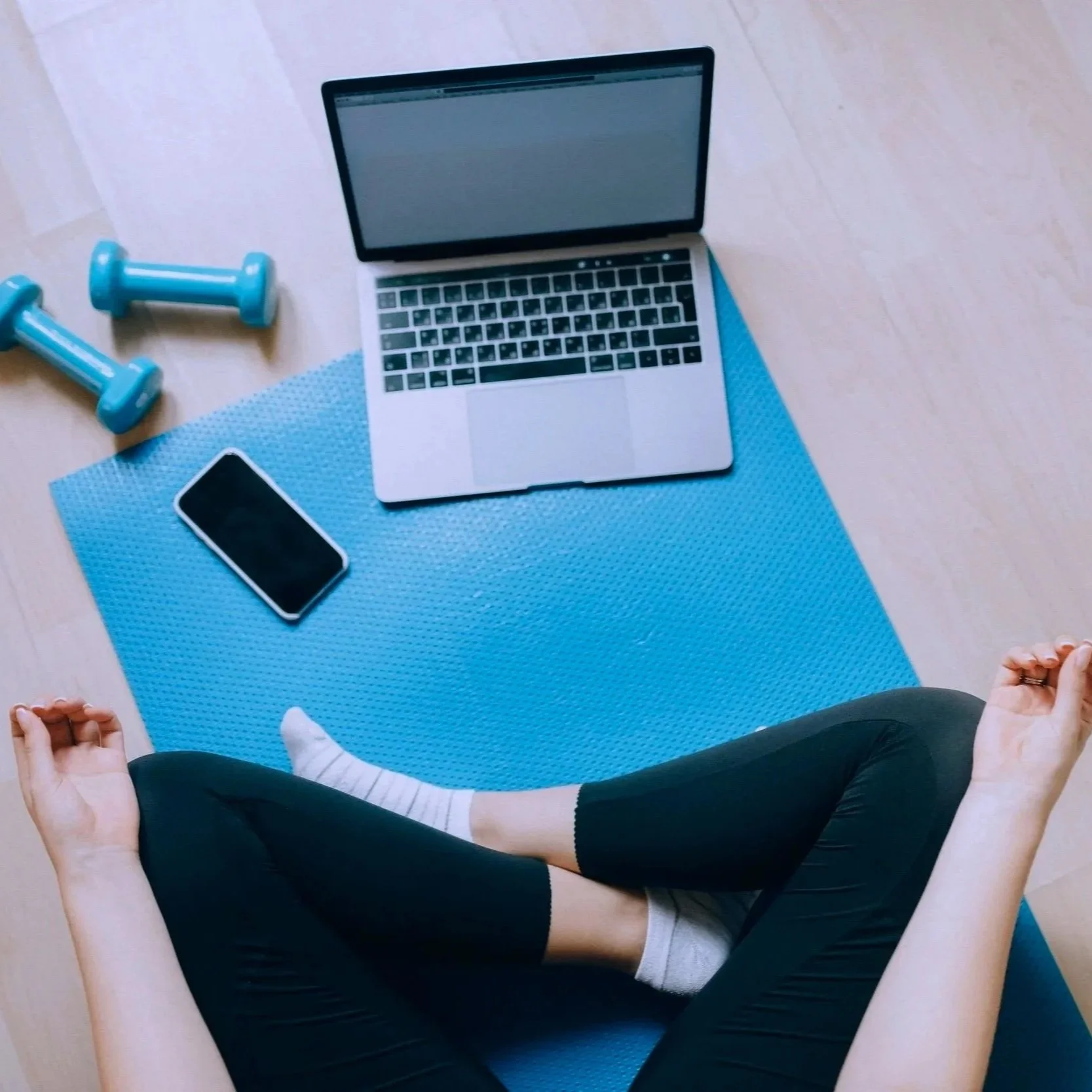 Person sitting on a blue yoga mat with crossing legs, workout equipment, a laptop, and a smartphone on the mat.