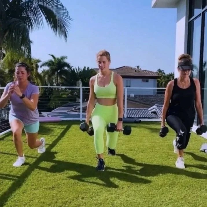 Three women exercising outdoors on a grassy area, lifting dumbbells, with houses and palm trees in the background.