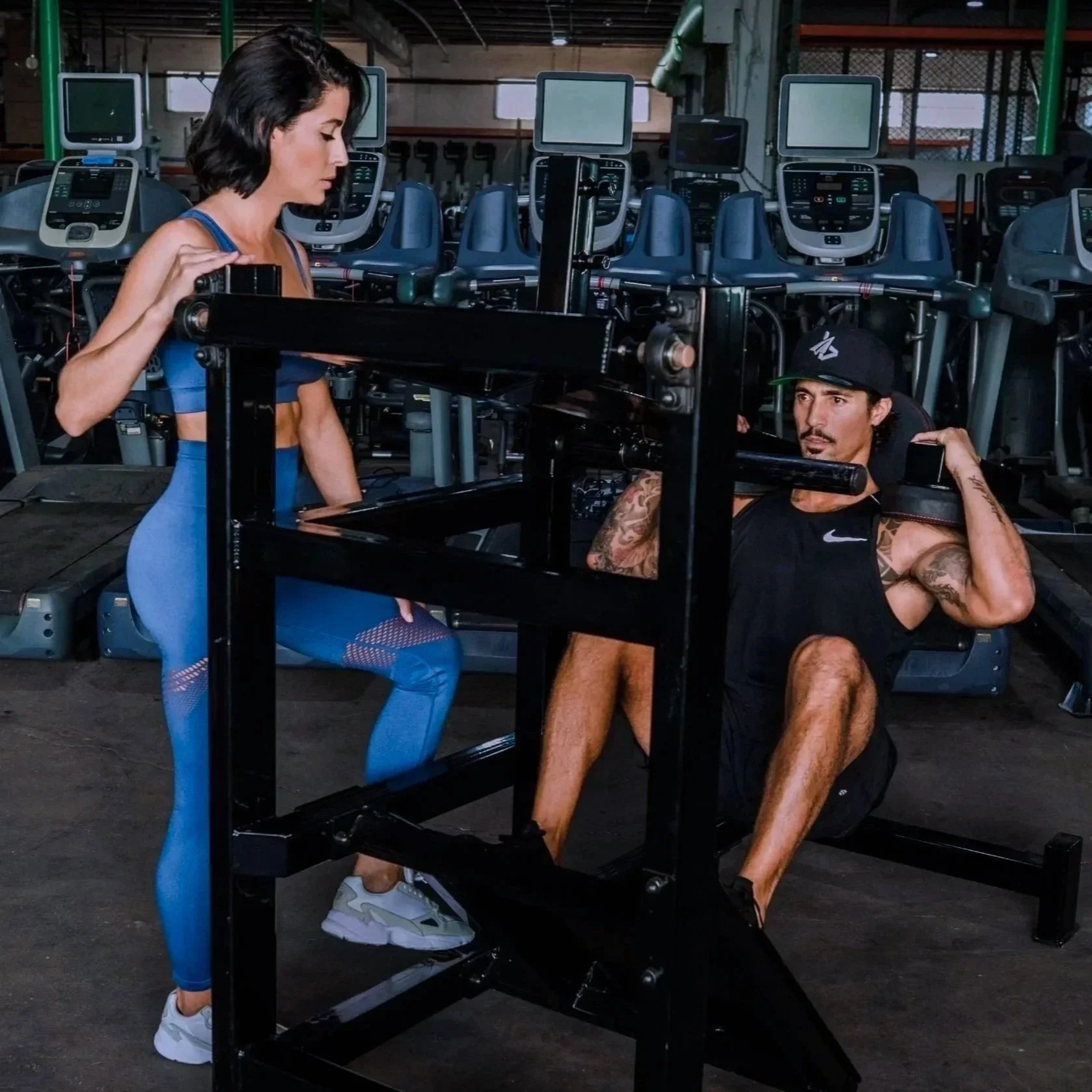 A woman in blue workout clothes instructs a man in black exercise clothes during a workout session at a gym, with treadmills in the background.