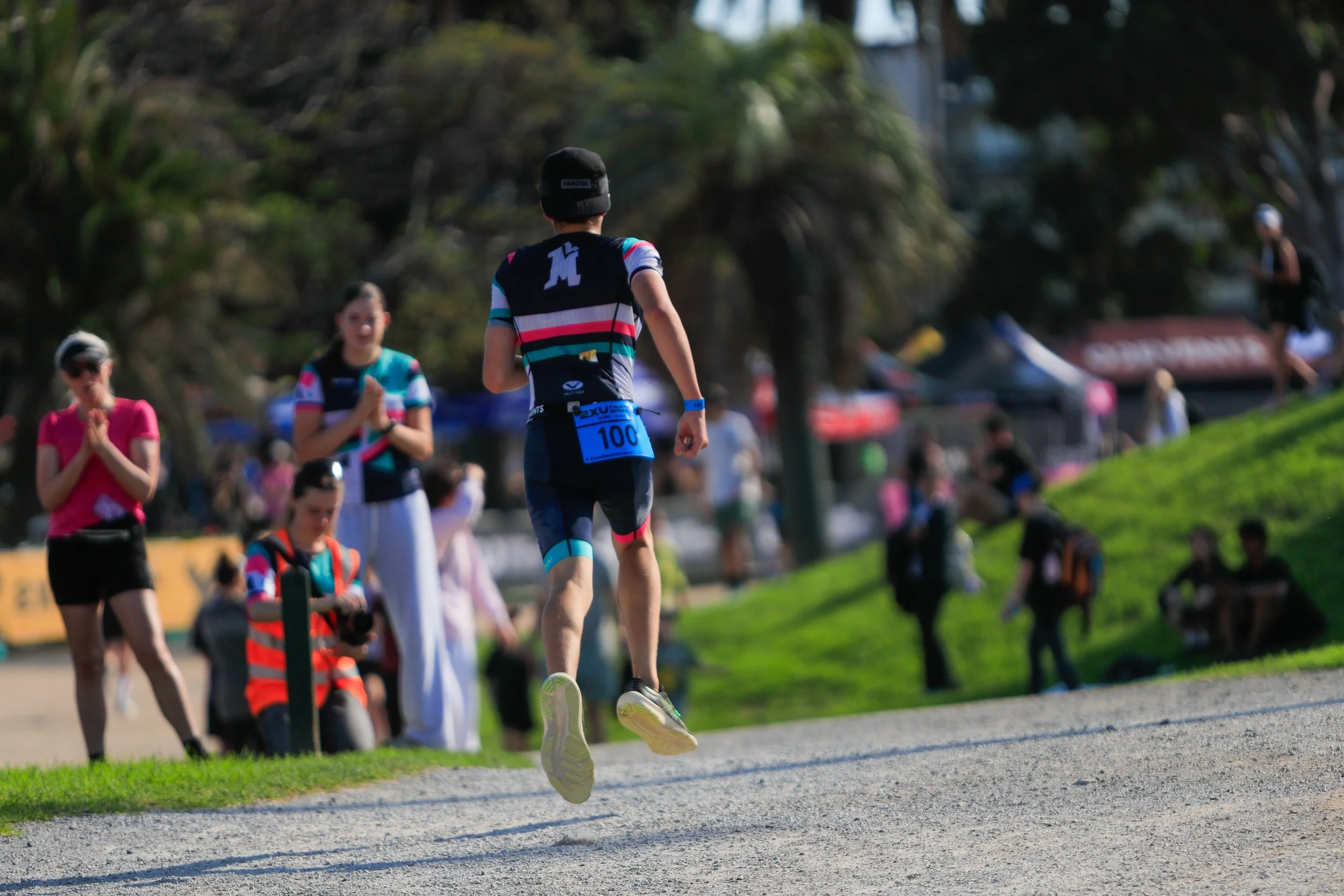 A person participating in a race, captured mid-air while running, in a park with several other people and trees in the background.
