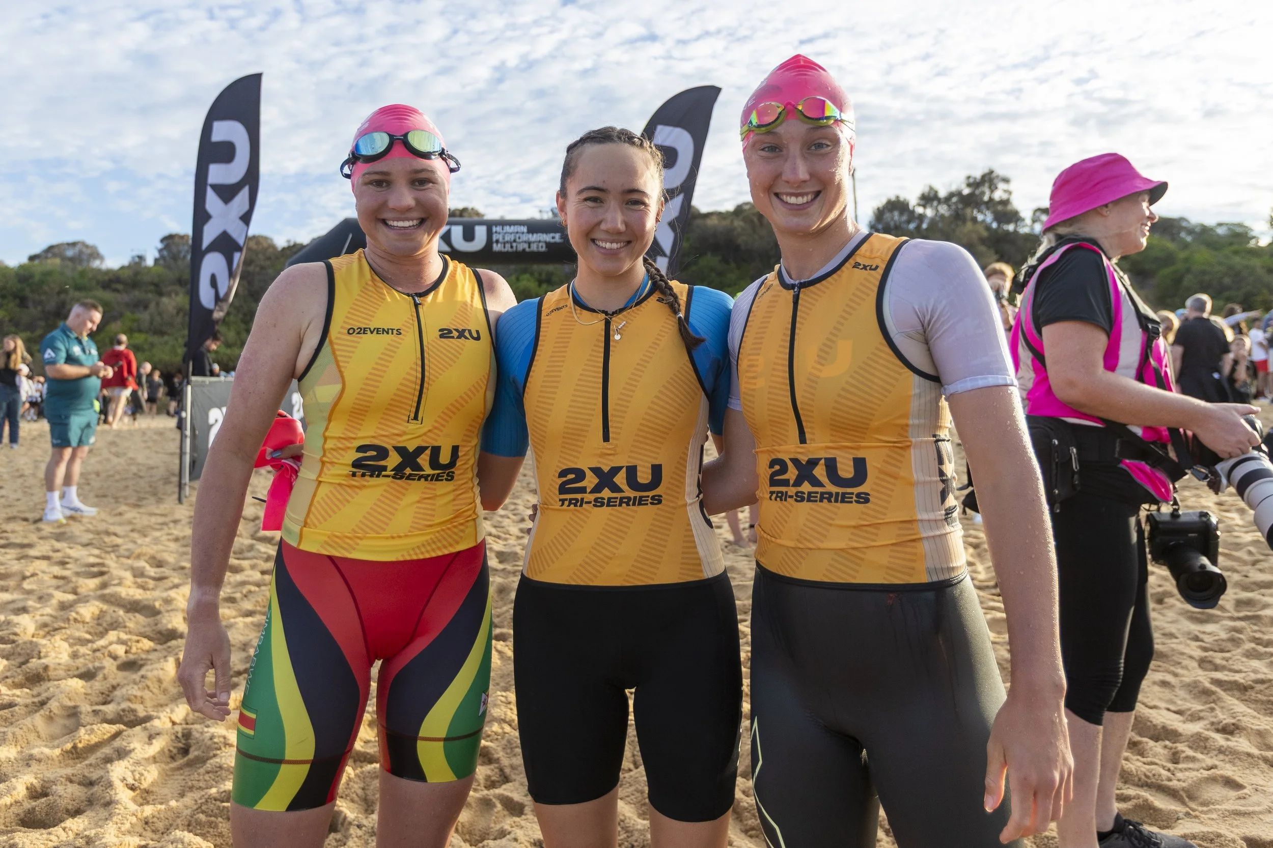 Four smiling female triathlon athletes in colorful sportswear, including yellow, black, and rainbow leggings, standing on sandy ground after a race, with flags and other participants in the background under a cloudy sky.