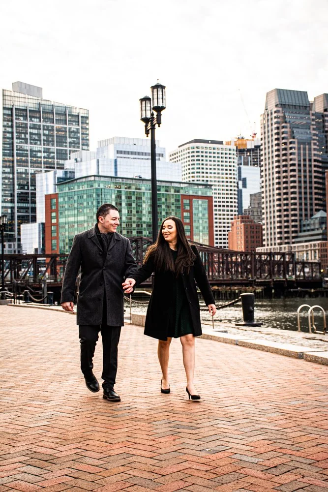 A man and woman walking hand-in-hand along a brick pathway in an urban area with tall buildings, a body of water, and a bridge in the background.