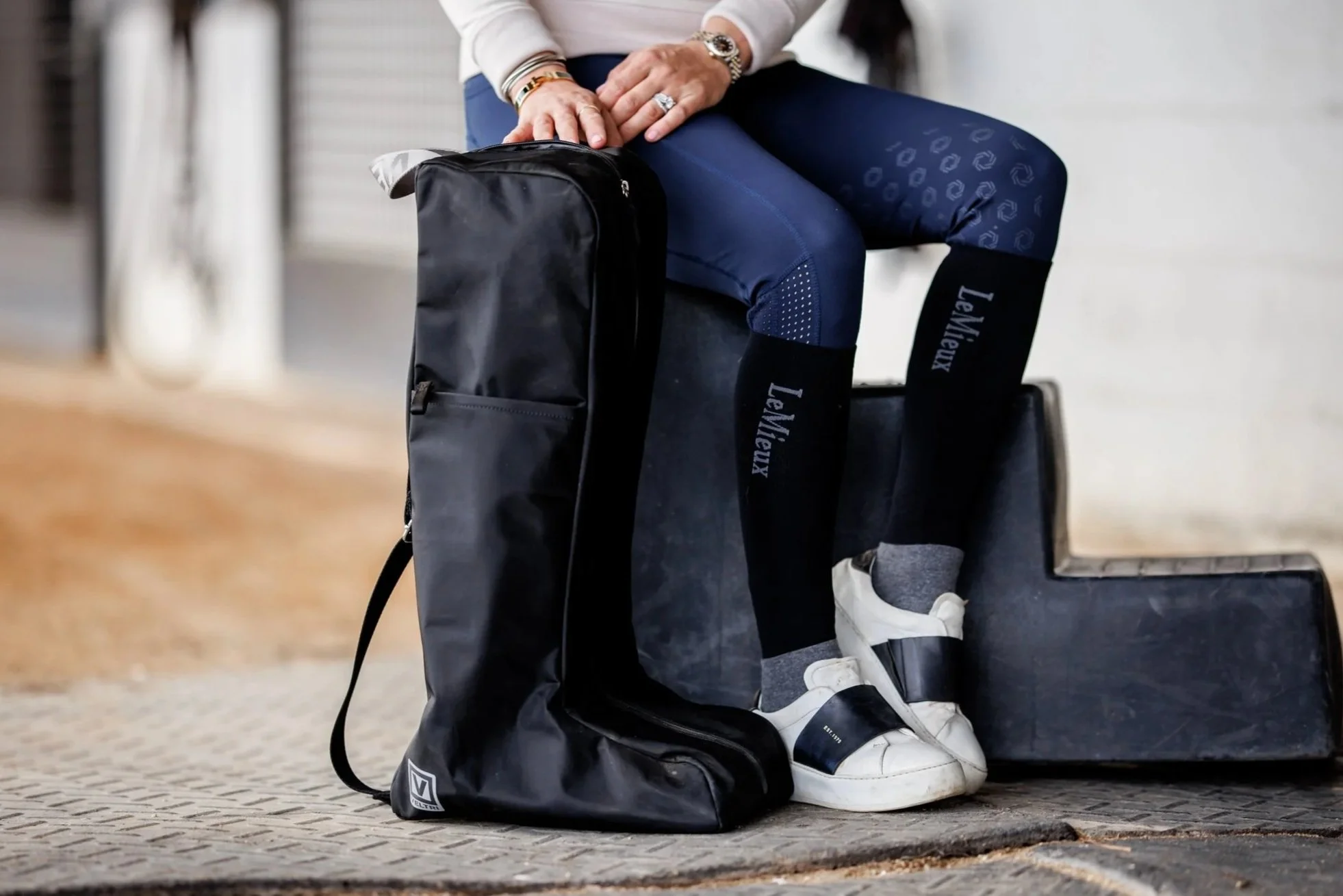 Person sitting on a bench with equestrian equipment including a boot bag, wearing navy blue breeches, white sneakers, and socks with the brand name 'LeMieux', in a barn setting.