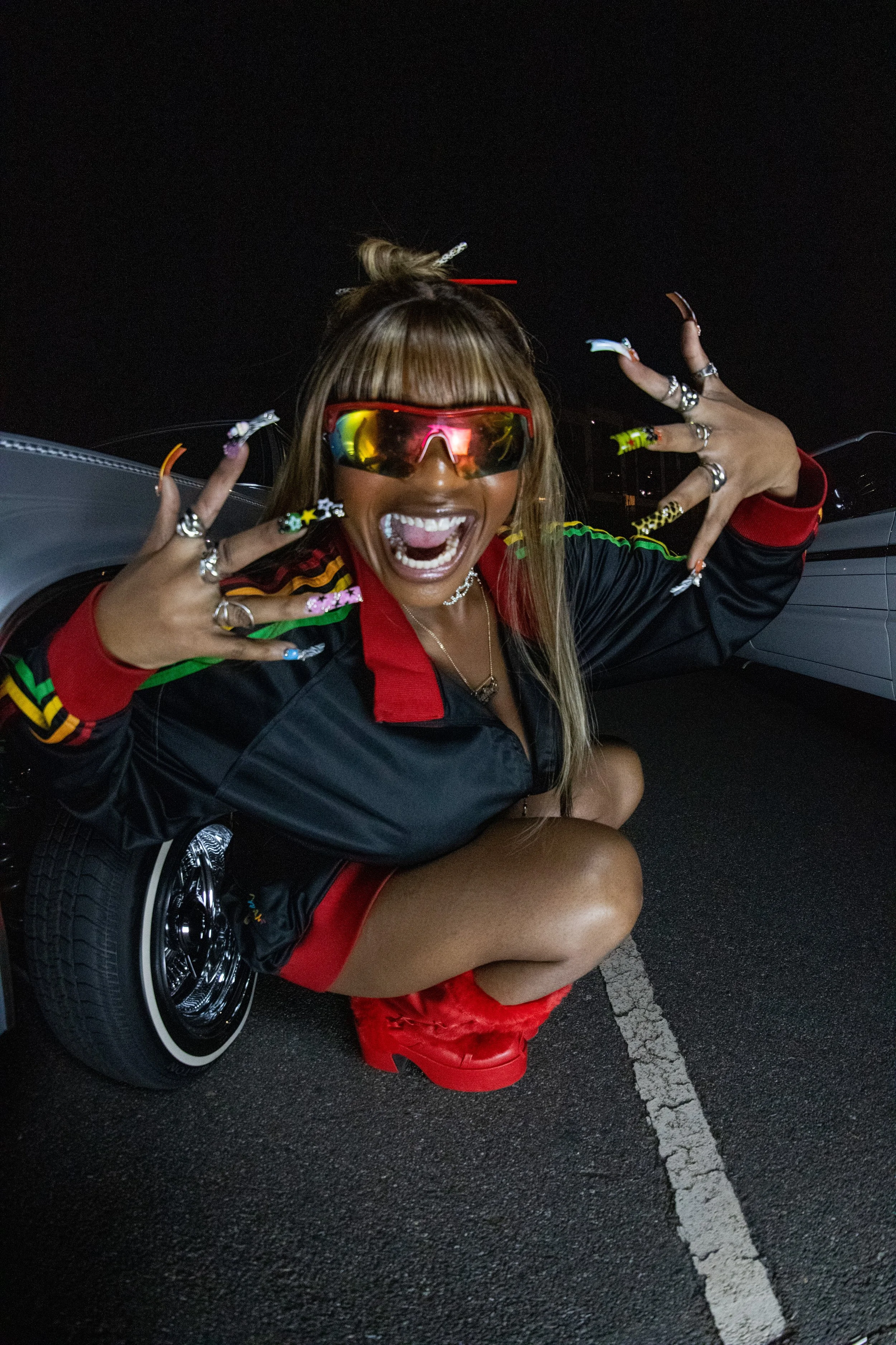 A woman with long blonde hair, wearing reflective sunglasses, colorful nail art, and red boots, squatting near a classic car at night, making expressive hand gestures and smiling widely.